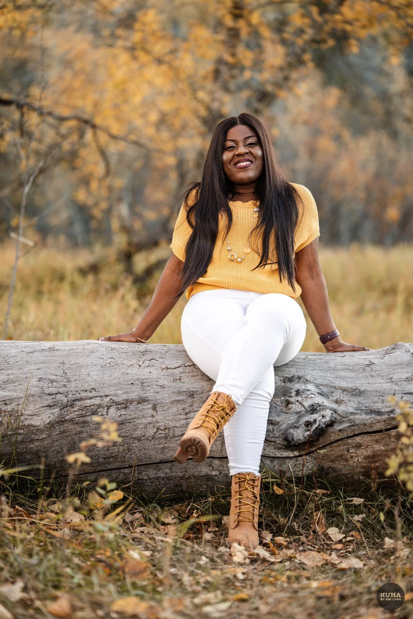 A woman sitting on a fallen log outdoors in a park or forested area with autumn foliage, wearing a yellow top, white pants, and tan boots, smiling and looking at the camera.