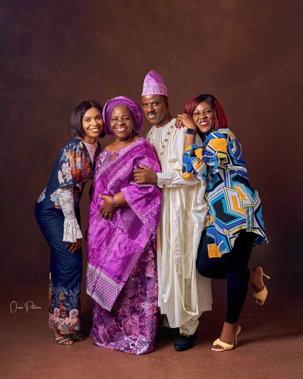 Four people, two women and two men, dressed in colorful traditional and modern African clothing, standing close and smiling at the camera against a brown background.