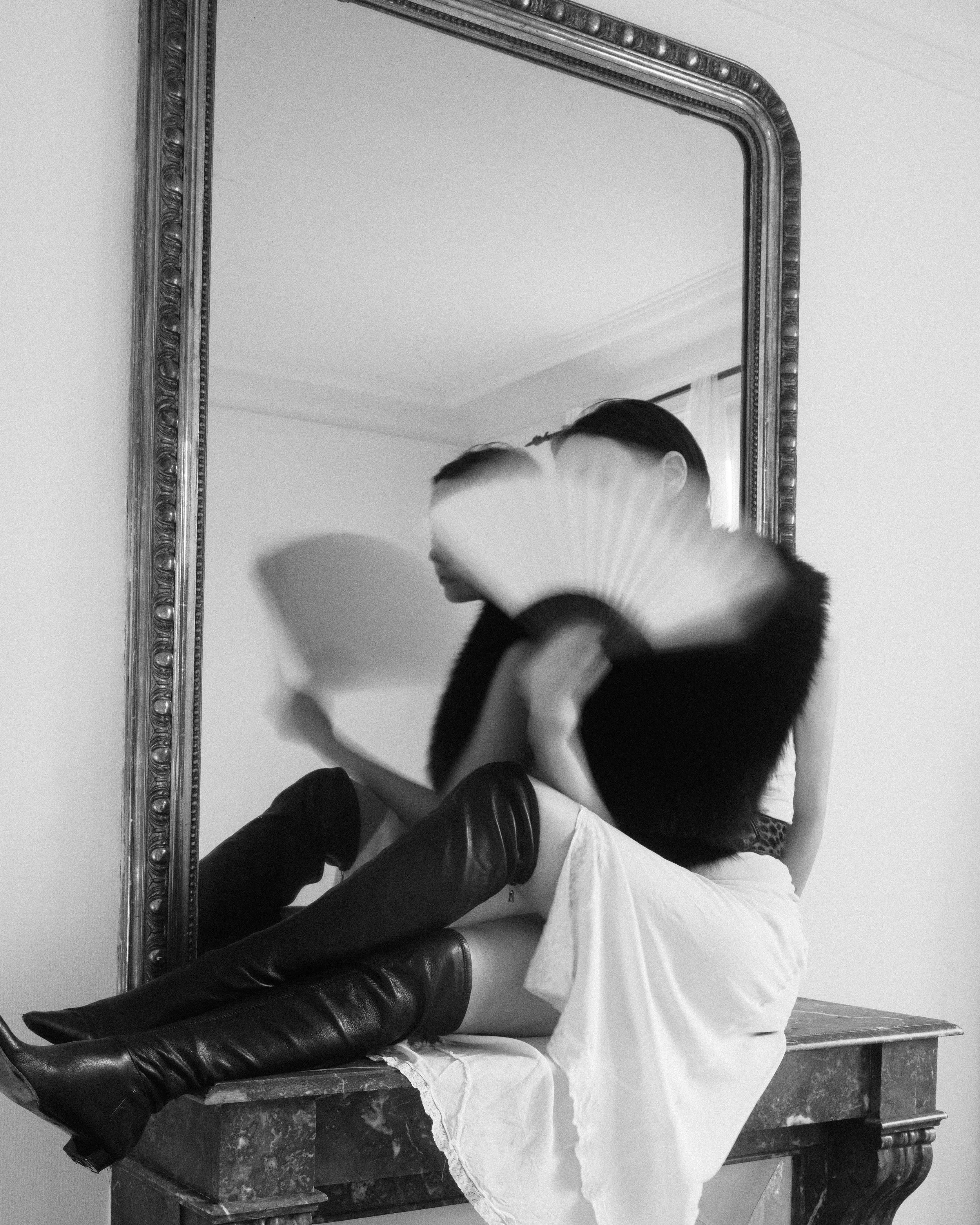 A black and white photo of Kayten Schmidt sitting on a marble mantle in front of a large ornate mirror, holding a fan, wearing thigh-high boots.