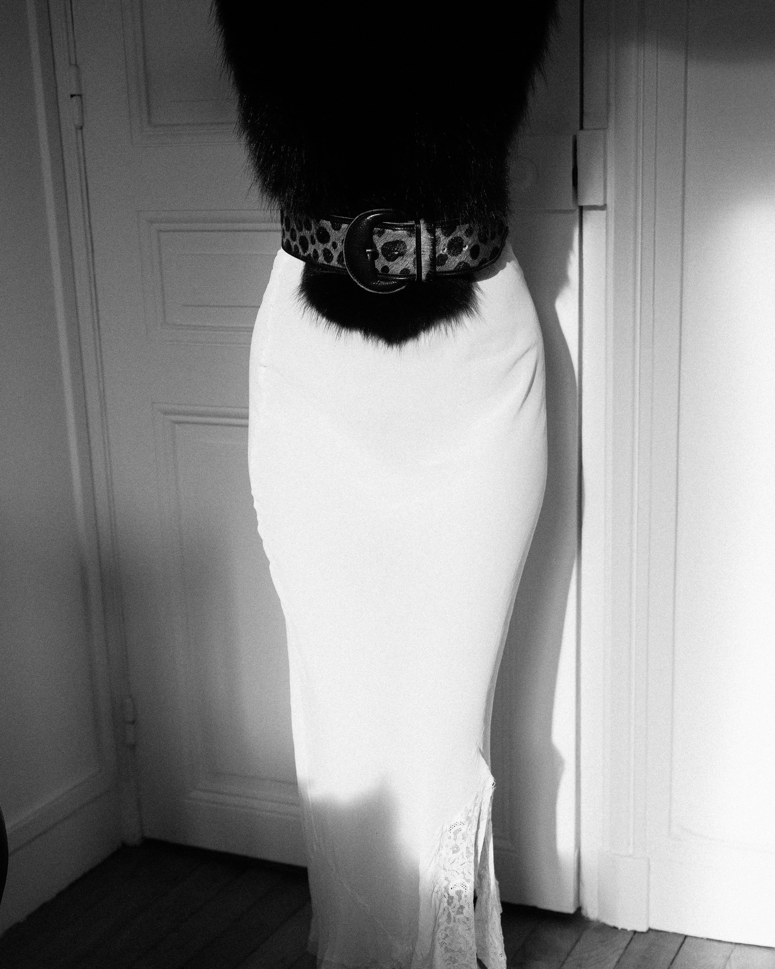 Kayten Schmidt wearing a long, white 1930s nightgown, and a leopard print belt, with the upper half of their body obscured.