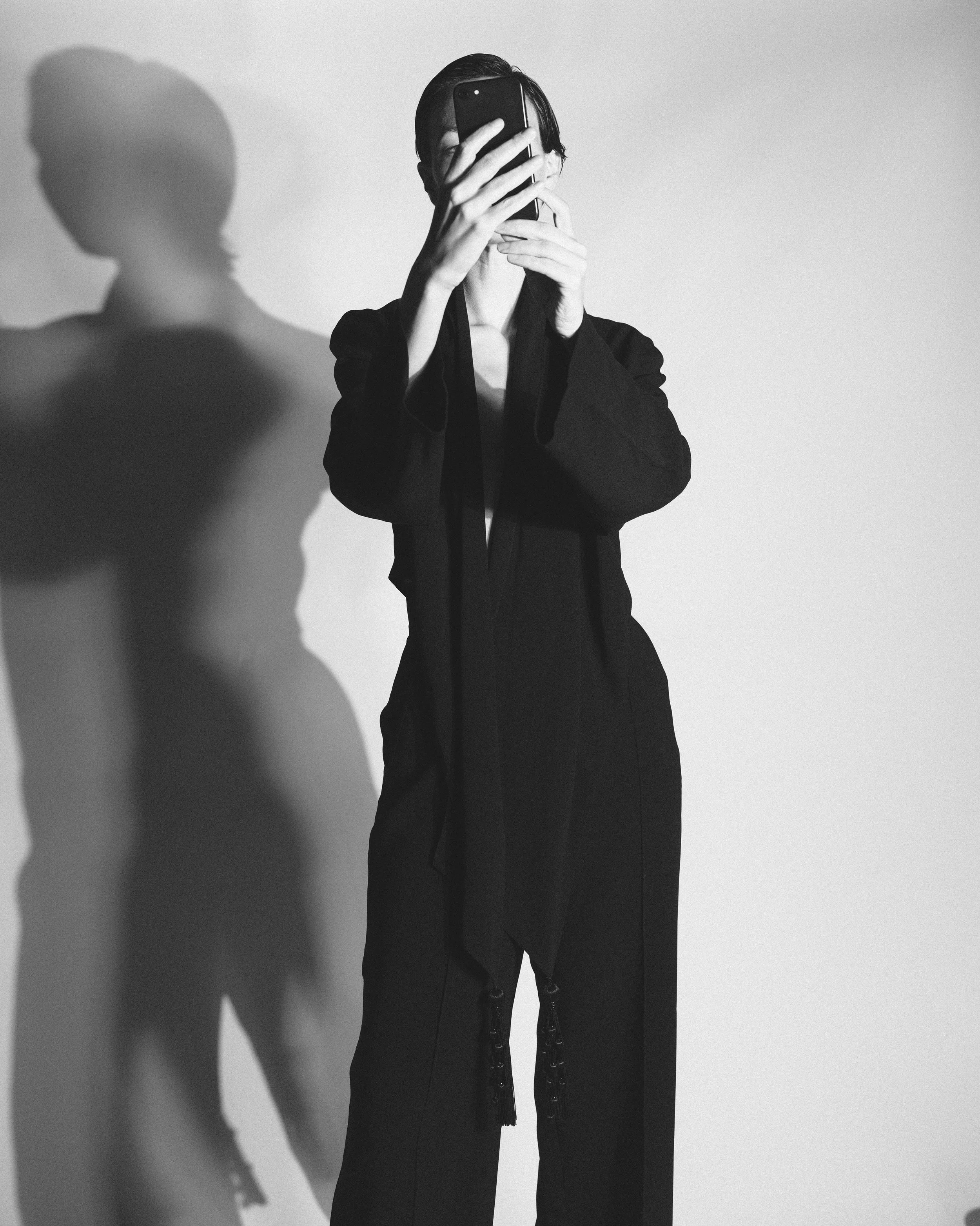 Kayten Schmidt in vintage Donna Karan taking a selfie with a smartphone. Their shadow is cast on the wall behind them.