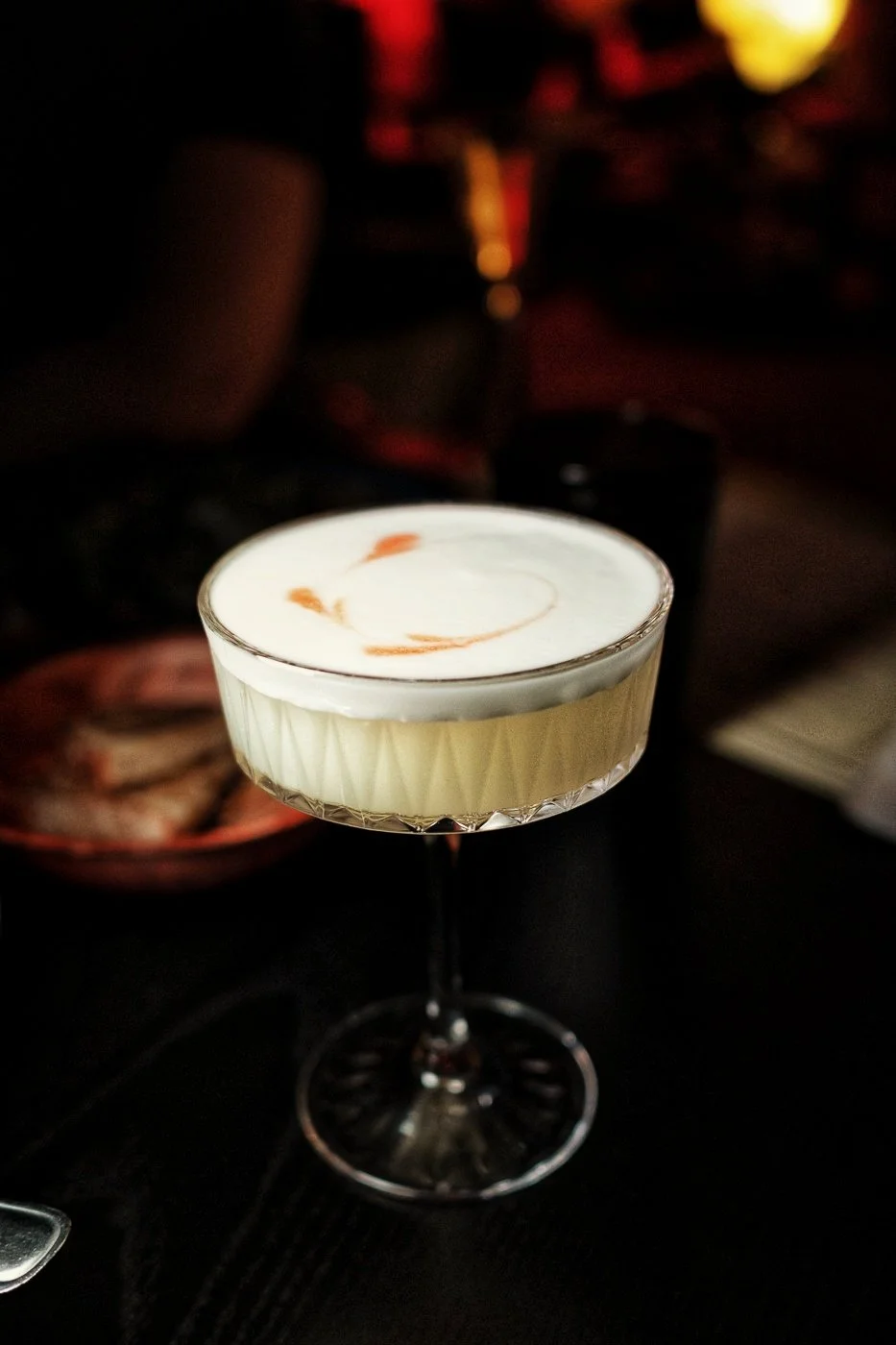A cocktail glass with a white creamy drink topped with a swirl of caramel or chocolate sauce, set against a dark background in a dimly lit setting, possibly a bar or restaurant.