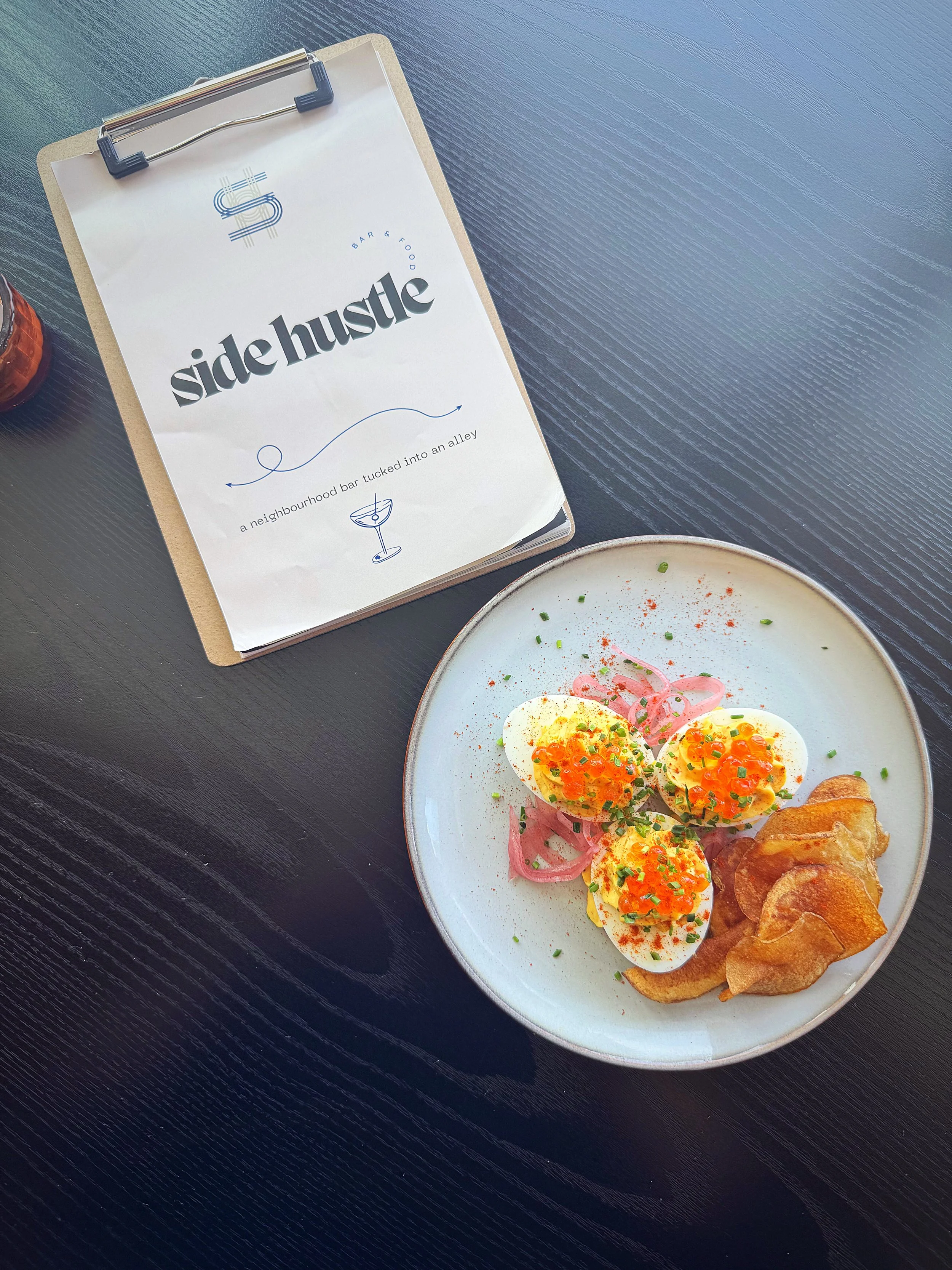 Smoked Trout Caviar - Devilled Eggs with Pickled Onion, Dill & Chives, garnished with pink pickled onions and accompanied by potato chips, next to a menu titled 'Side Hustle' on a dark table.