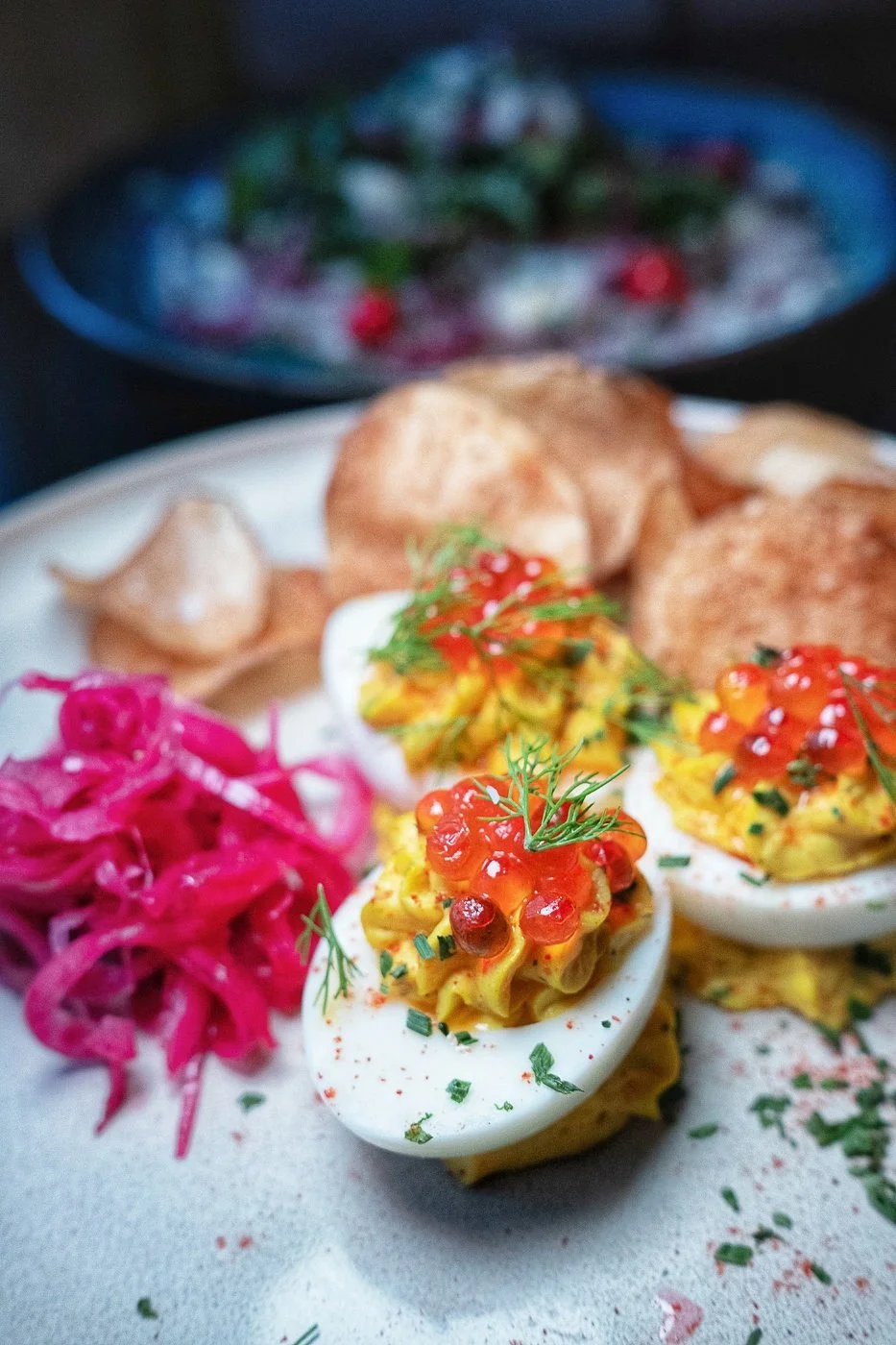 Deviled eggs topped with caviar and herbs, accompanied by pickled pink onions and fried potato chips, on a white plate.
