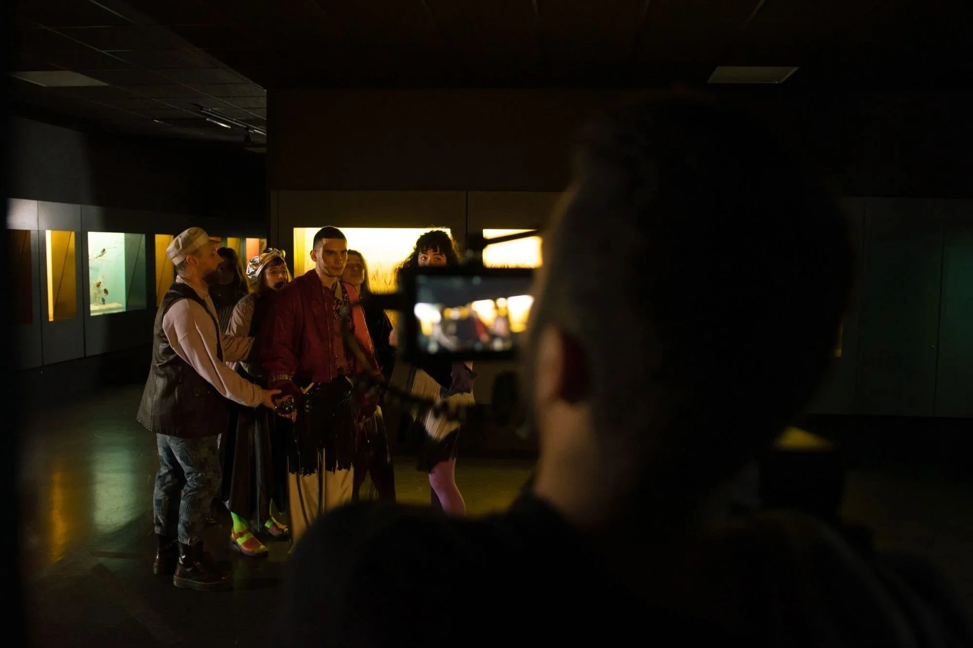 Group of people taking a photo or selfie in a dark room, with a line of illuminated display cases or exhibits in the background, possibly in a museum or exhibit space.