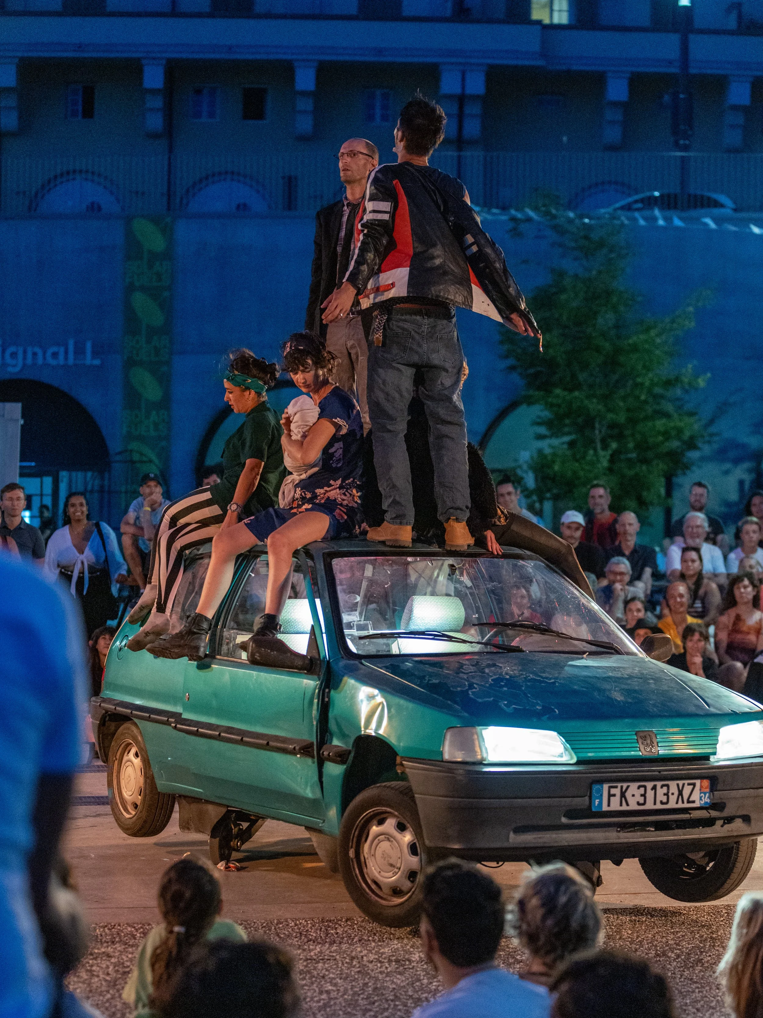 A green car is performing a stunt where it is lifted on its front wheels with people sitting on top of it. A crowd of spectators watches the scene in an urban area at dusk.