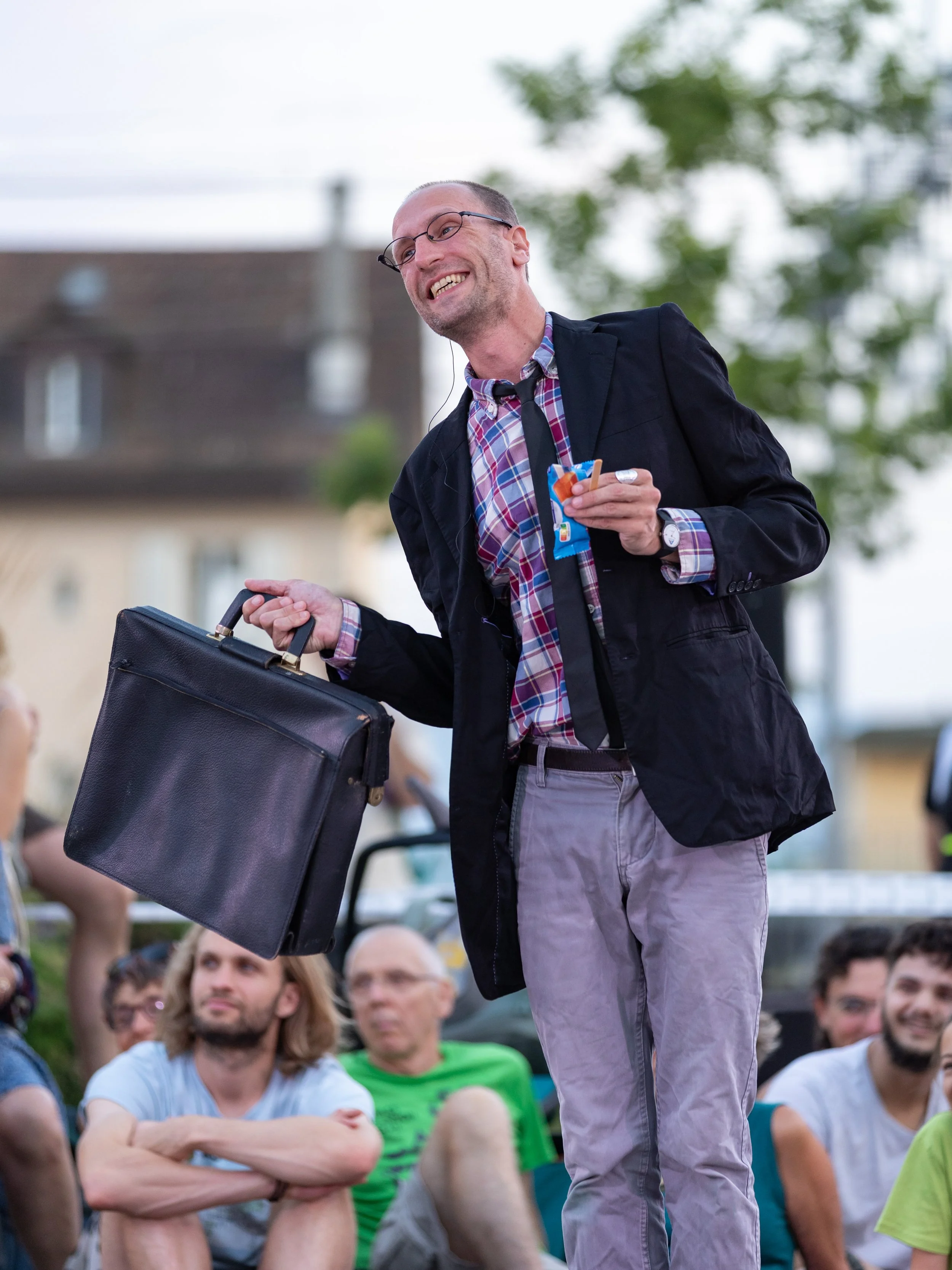 A man with glasses in a black jacket, plaid shirt, and light gray pants, smiling while holding a black briefcase in one hand and a snack in the other, standing in front of a seated audience outdoors.