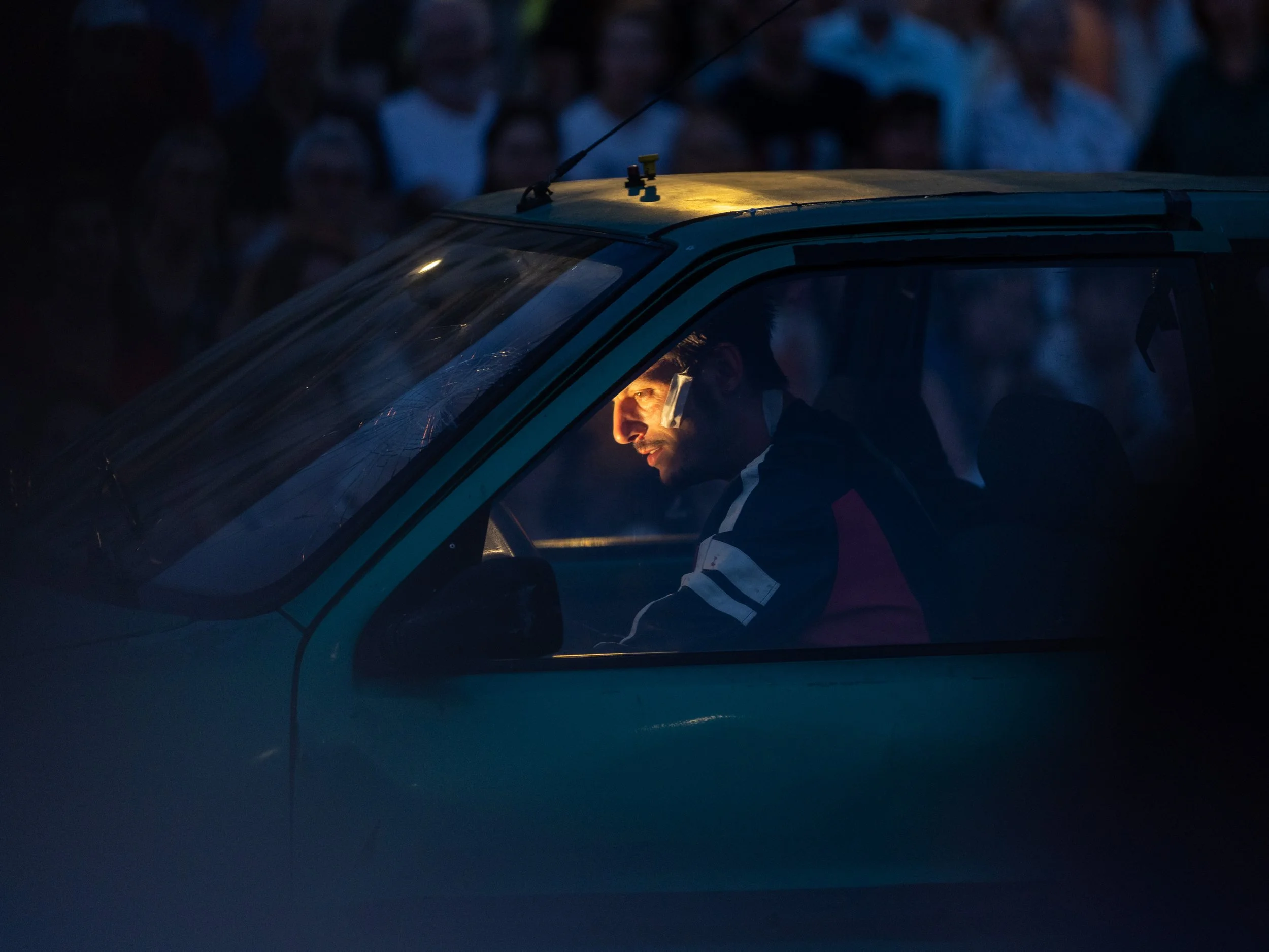 A man with light skin, wearing a jacket, sitting inside a blue vehicle, looking out the window with a serious expression. The vehicle's roof and part of the windshield are visible, with a dark background.