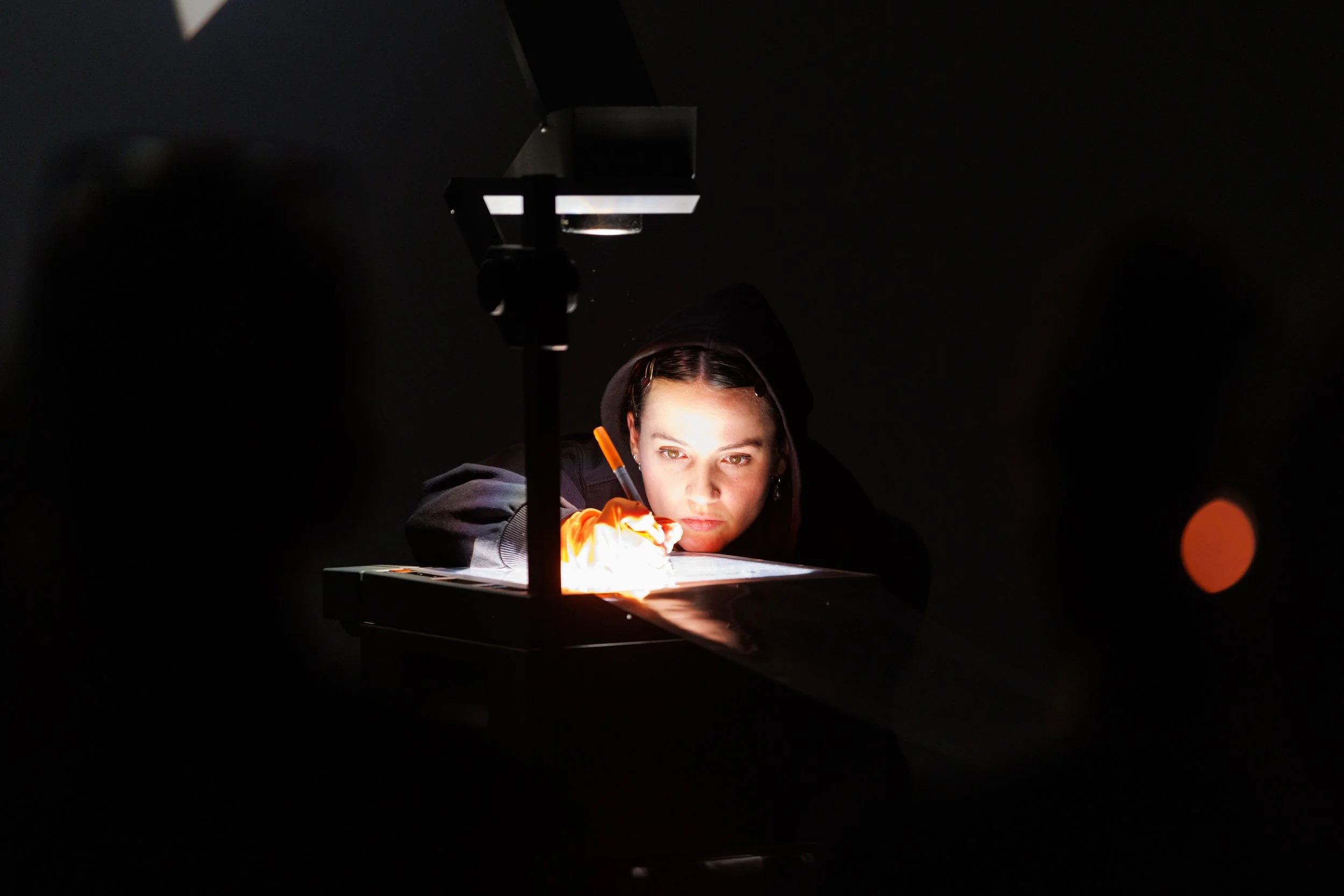 A person wearing a hoodie is working on a project under a focused light, with tools and materials on the table.