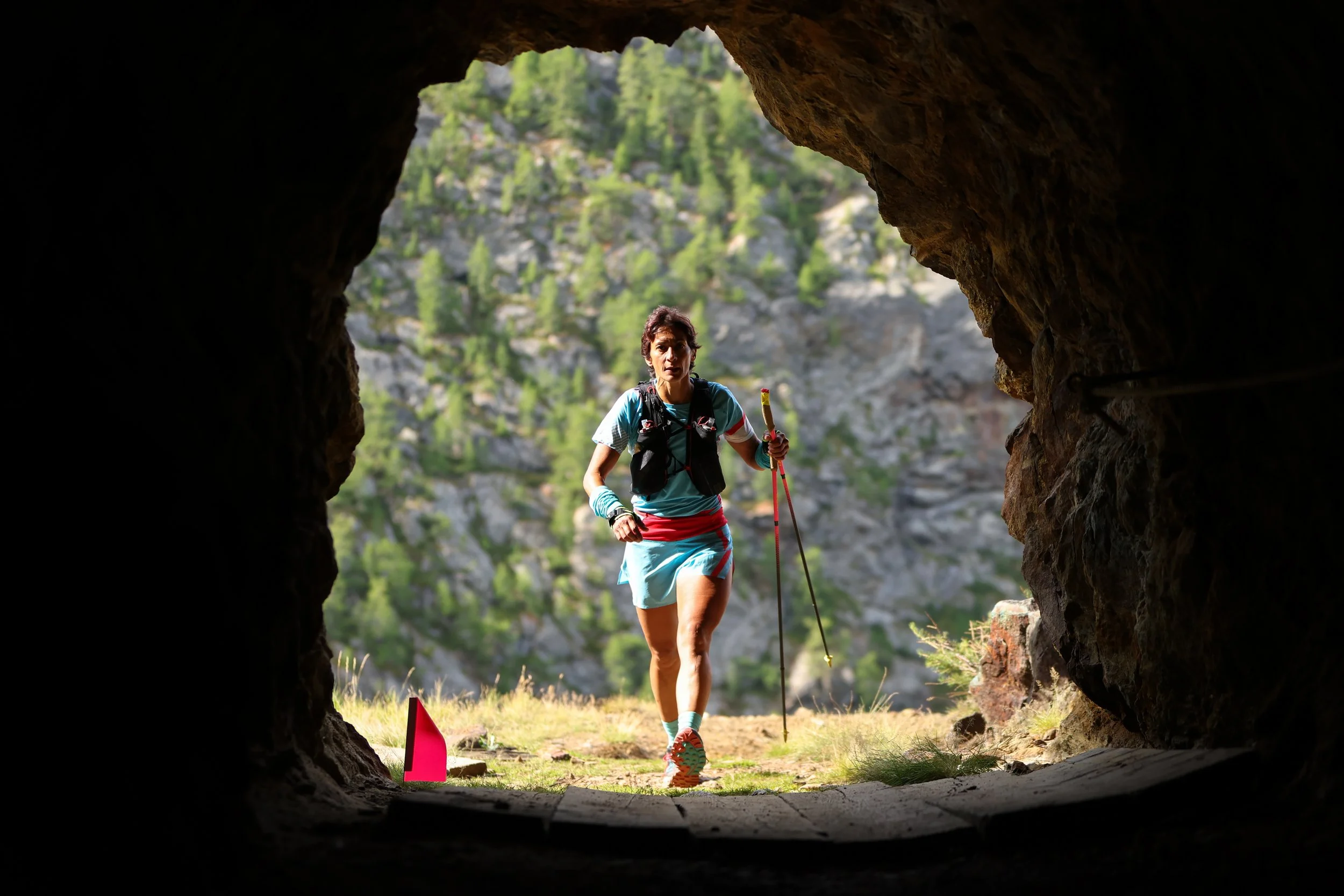 A woman hiking out of a dark cave, holding trekking poles, wearing athletic gear, with mountains and trees in the background.