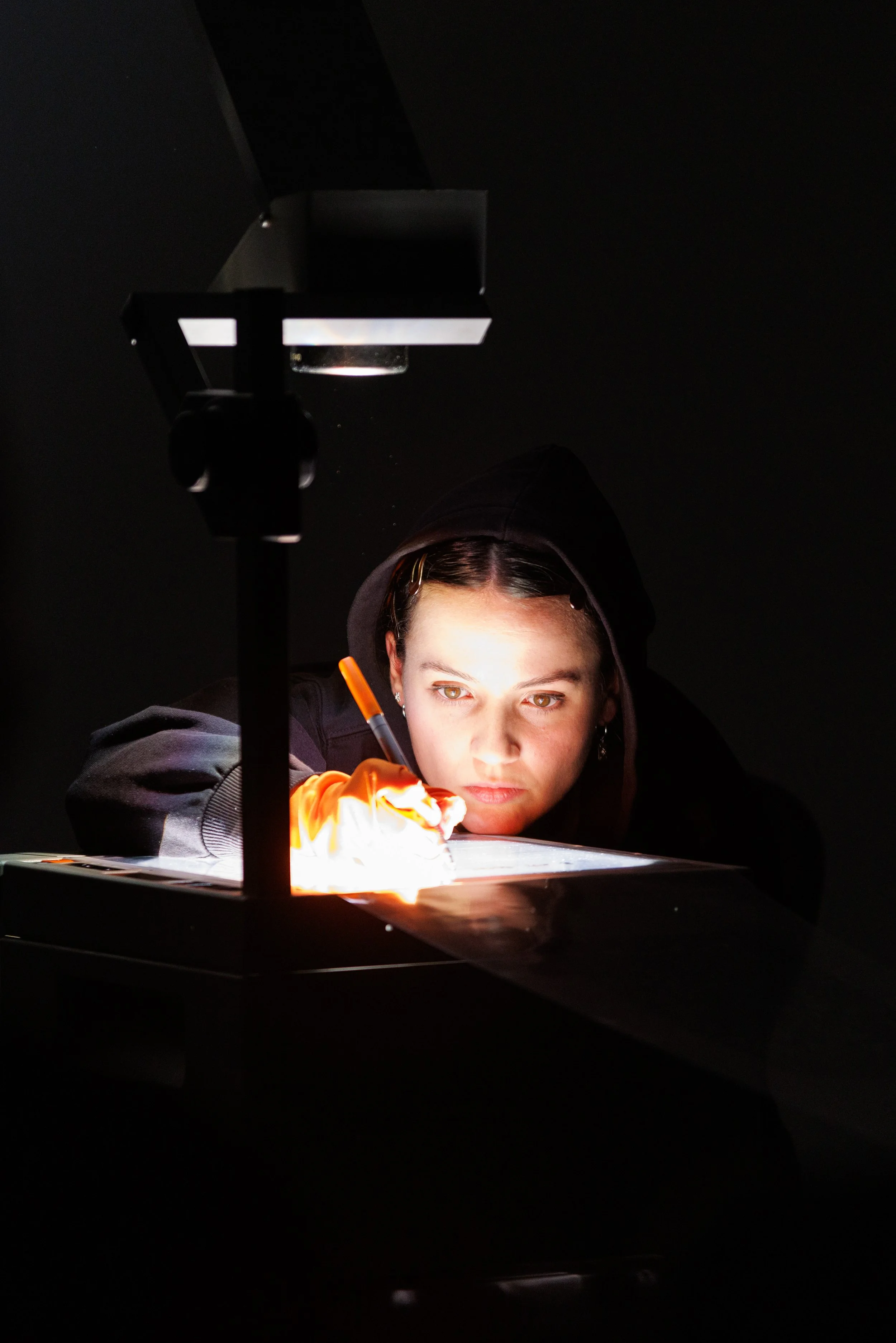 A person with light skin in a dark room, wearing a hoodie and earrings, is looking at a projection of a bright, swirling light, possibly a flame or a light source, on a surface in front of them.
