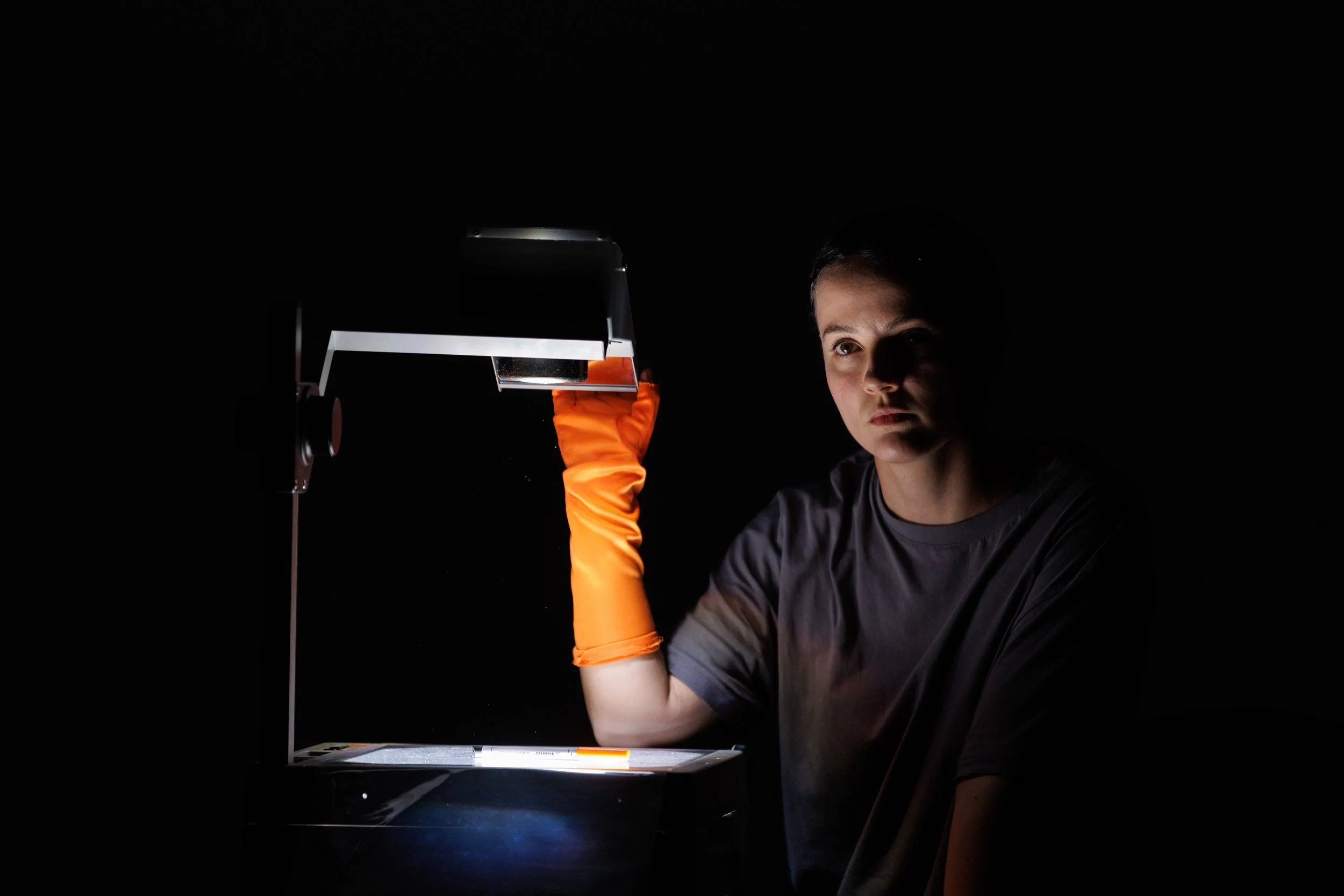 A young person with short dark hair wearing a gray shirt and orange gloves, examining something under a light in a dark room.