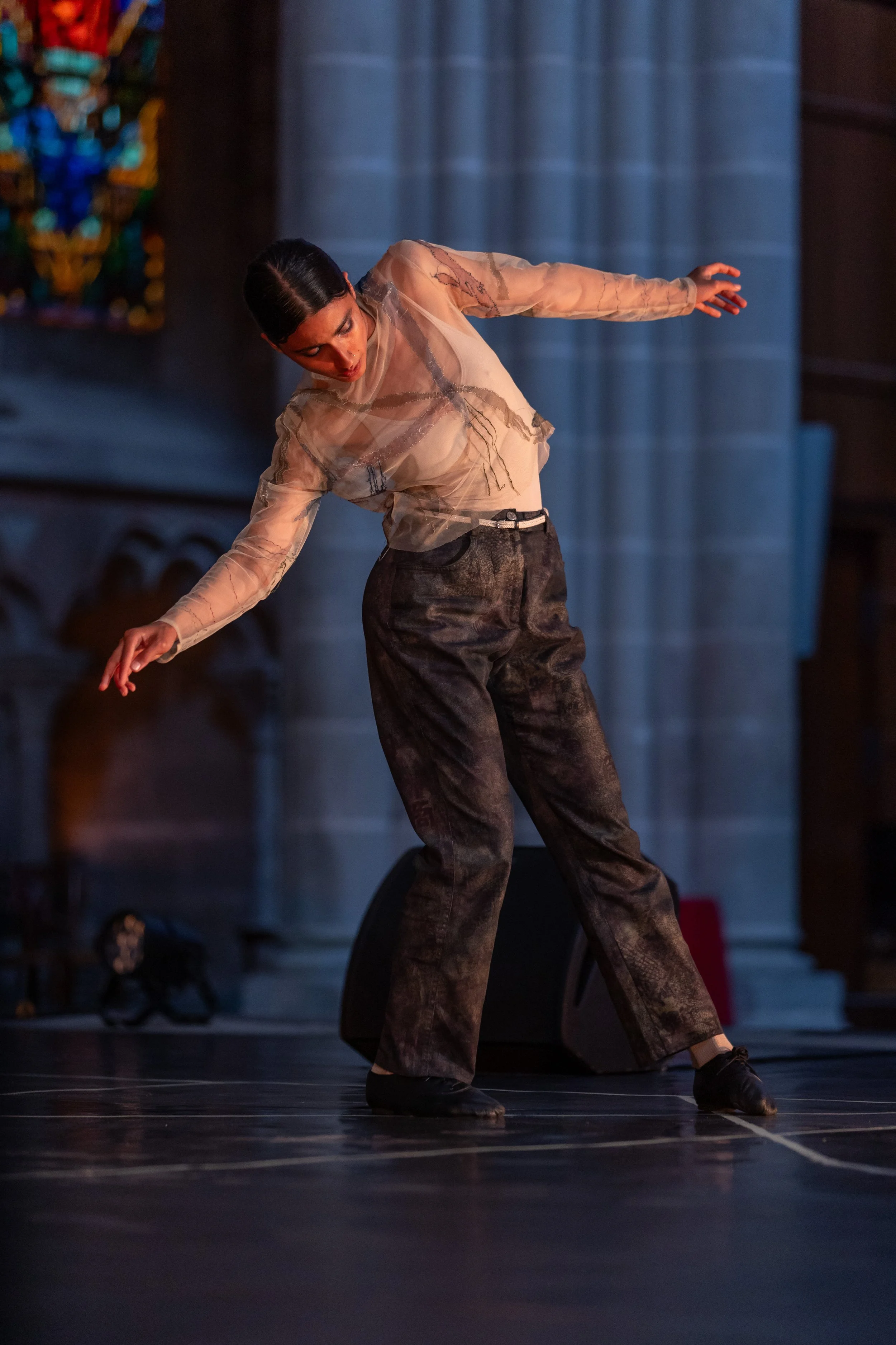 A female dancer in sheer, embroidered top and loose trousers performs on stage with a background of stone-like columns and stained glass window.