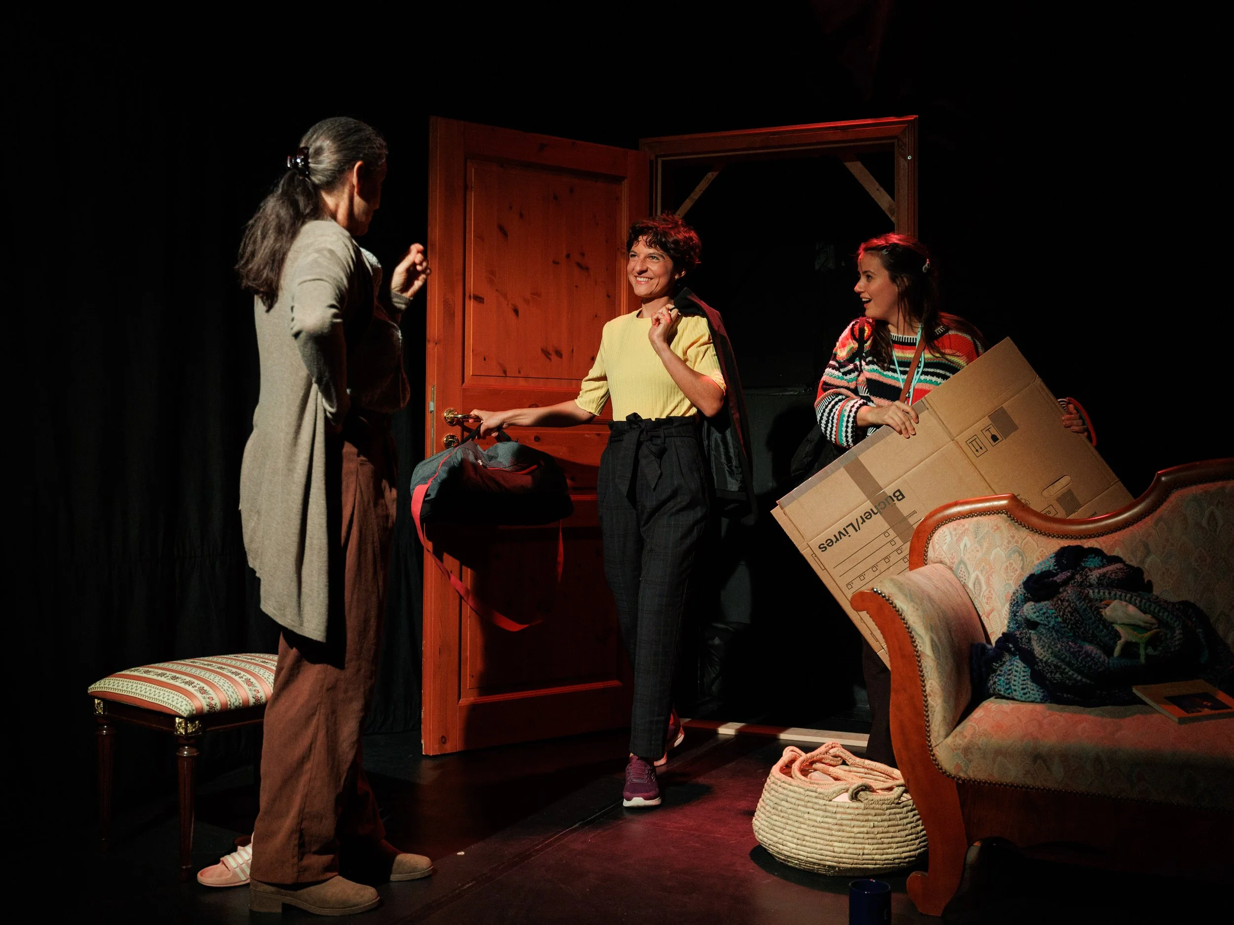 Three women on a stage, two standing and one smiling, with a wooden door in the background. The woman on the right holds a cardboard box, and other items such as a backpack and basket are nearby.