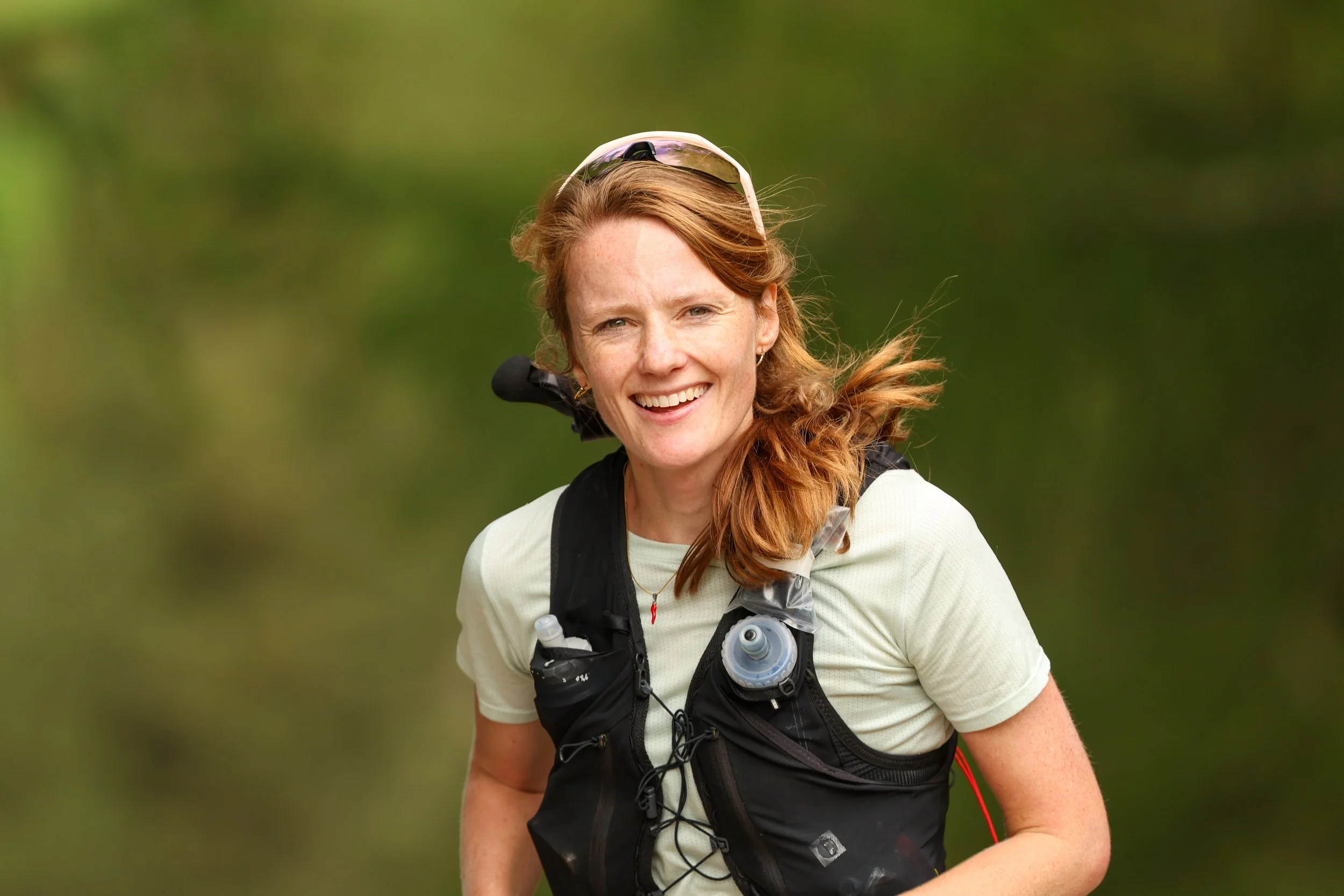 A smiling woman with red hair, wearing a white shirt, sunglasses on her head, and a black running vest with water bottles, outdoors on a green background.