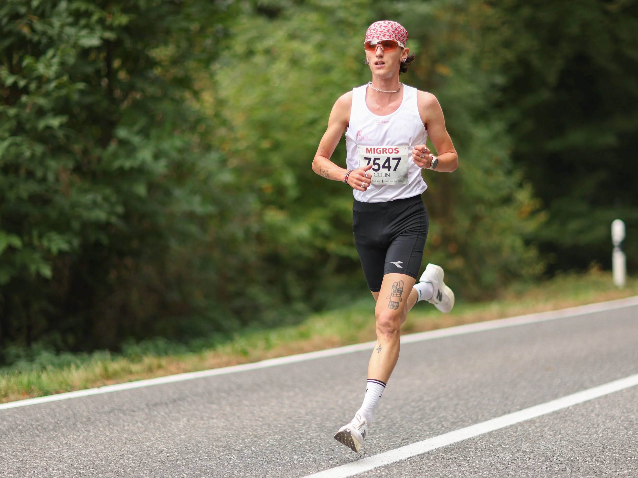A female runner wearing sunglasses, a headscarf, a white tank top with race number 7547, black shorts, and white socks running along a paved road surrounded by green trees.