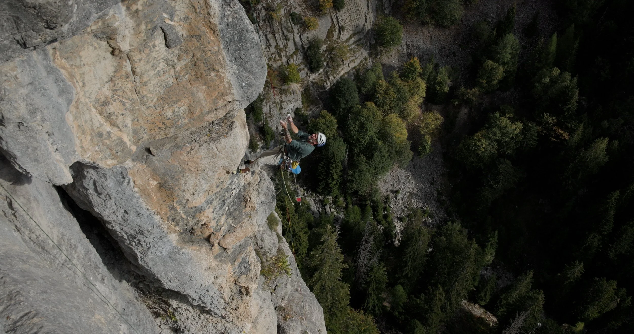 A person climbing a rock face with safety gear in a mountainous area surrounded by trees.