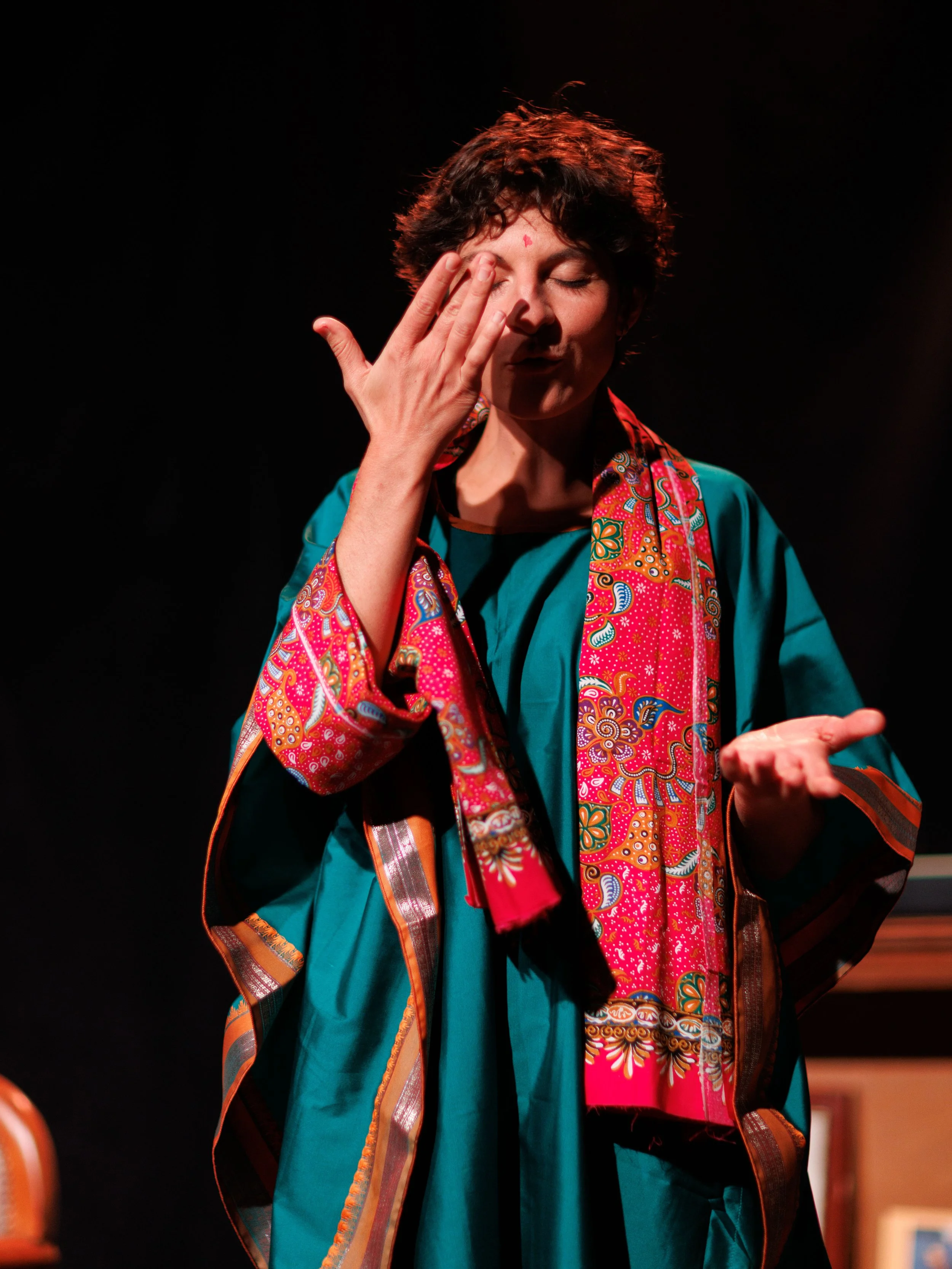 A woman with short curly hair and closed eyes touching her face with her left hand. She is wearing a colorful scarf and teal clothing, standing against a dark background.