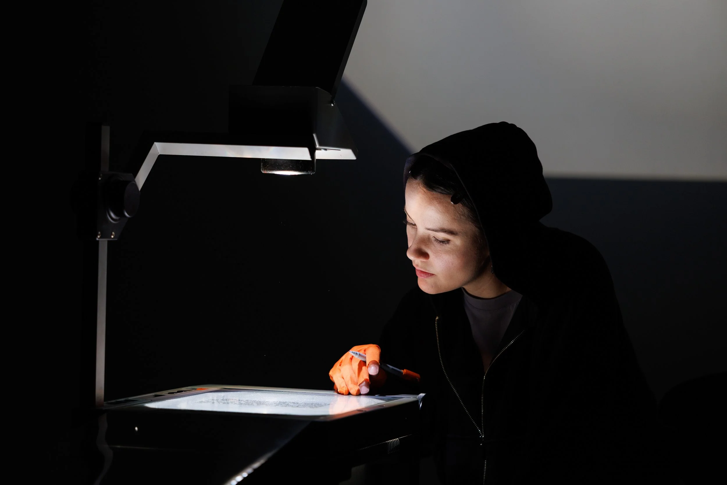 A person wearing a black hoodie and orange gloves is working with a small robotic or electronic device in a dark environment, illuminated by a focused light source.