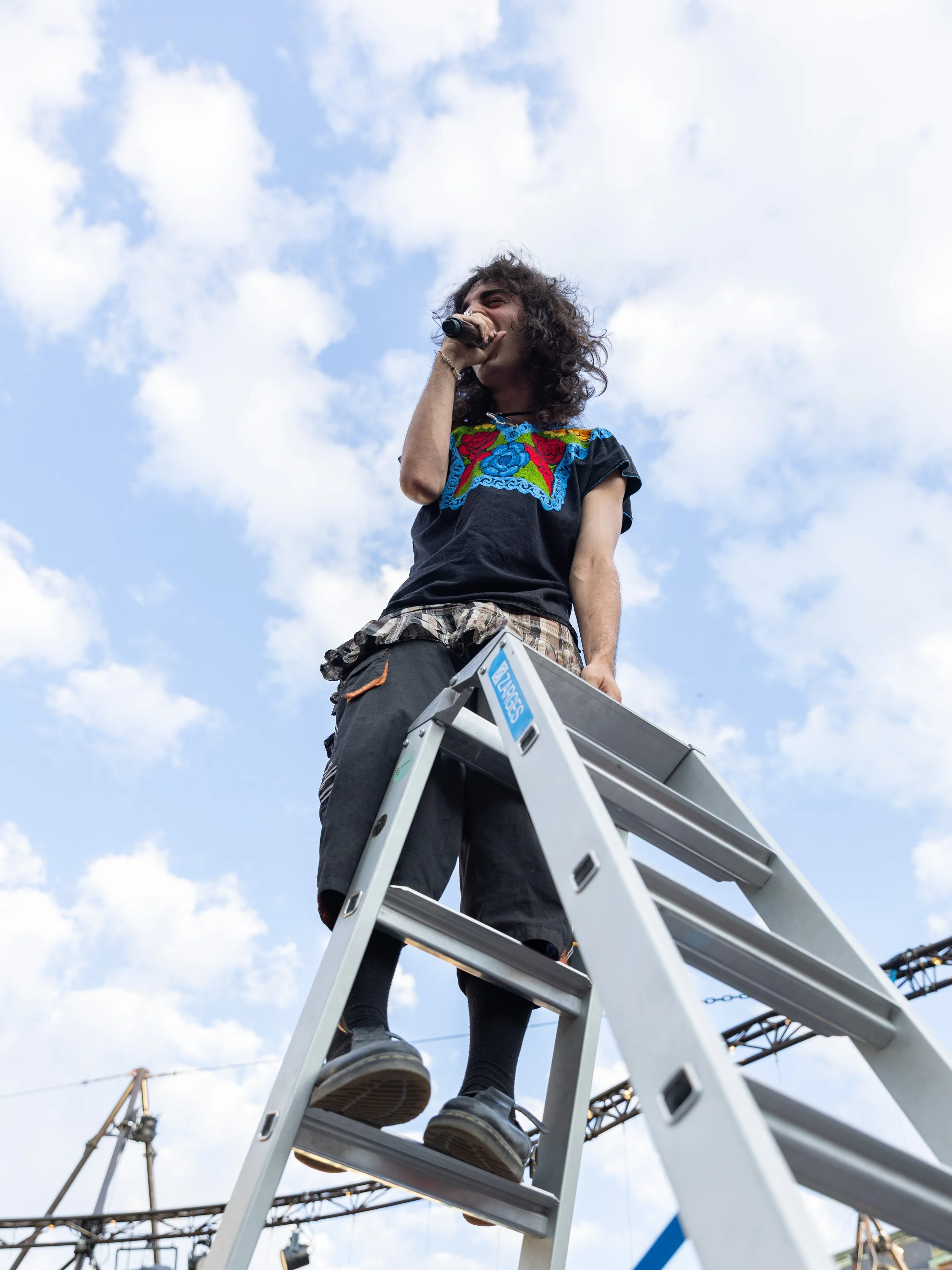 Person singing into a microphone on an outdoor stage, standing on a ladder, with construction cranes and a partly cloudy sky in the background.