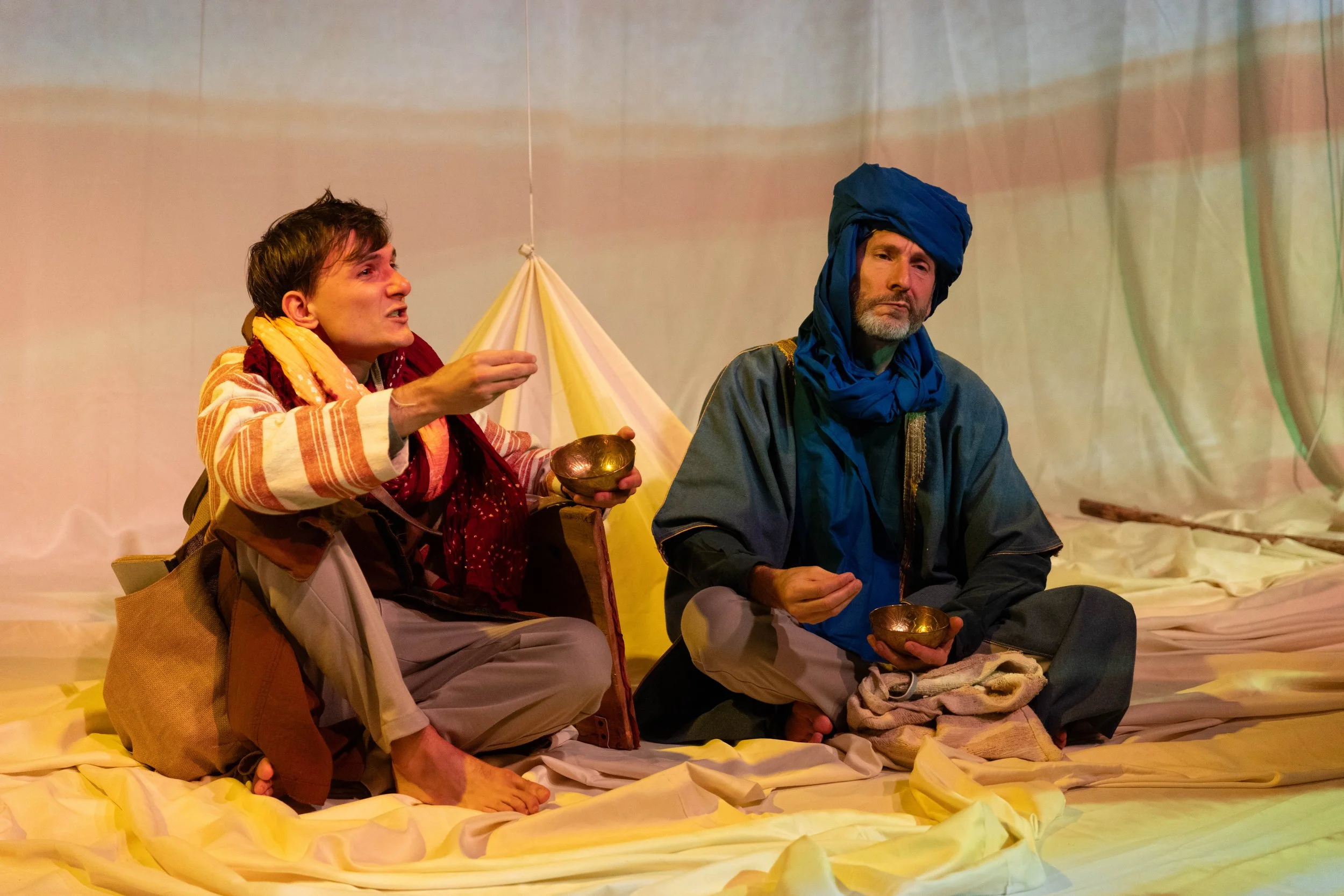 Two men sit on yellow fabric, dressed in traditional Middle Eastern attire, with one wearing a blue turban and the other holding a small bowl, engaging in a conversation on a stage with a light backdrop.