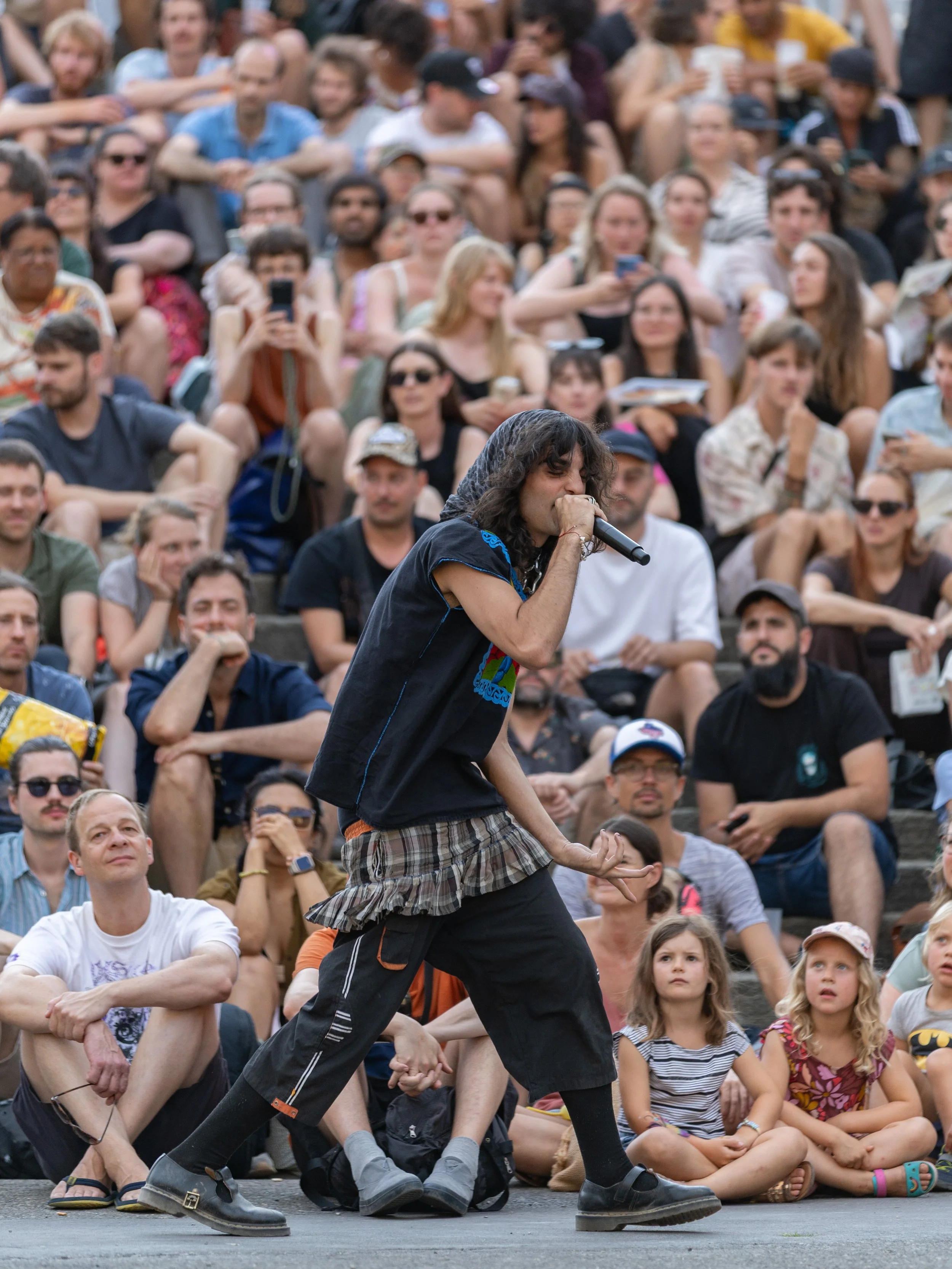 A man with long, dark hair wearing a black outfit and patterned shorts is performing in front of a large seated crowd at an outdoor event, holding hands with a woman.
