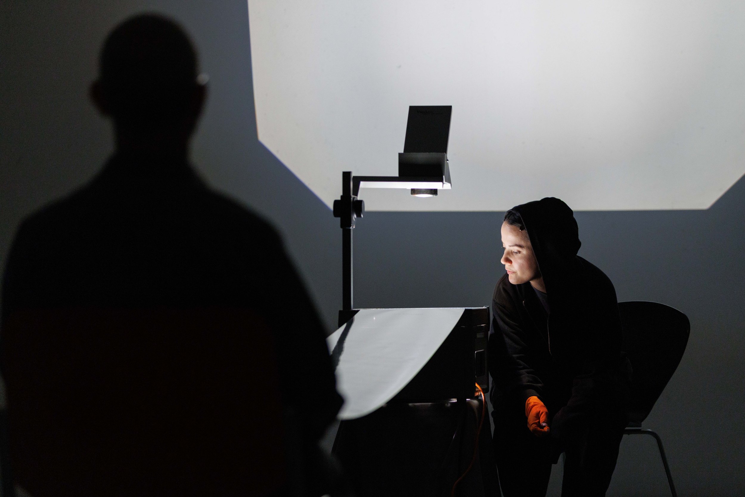A person wearing a black hoodie and orange gloves leaning towards a table with a bright light shining on their face, with a black laptop and a lamp in the background. Another person's silhouette is visible in the foreground.