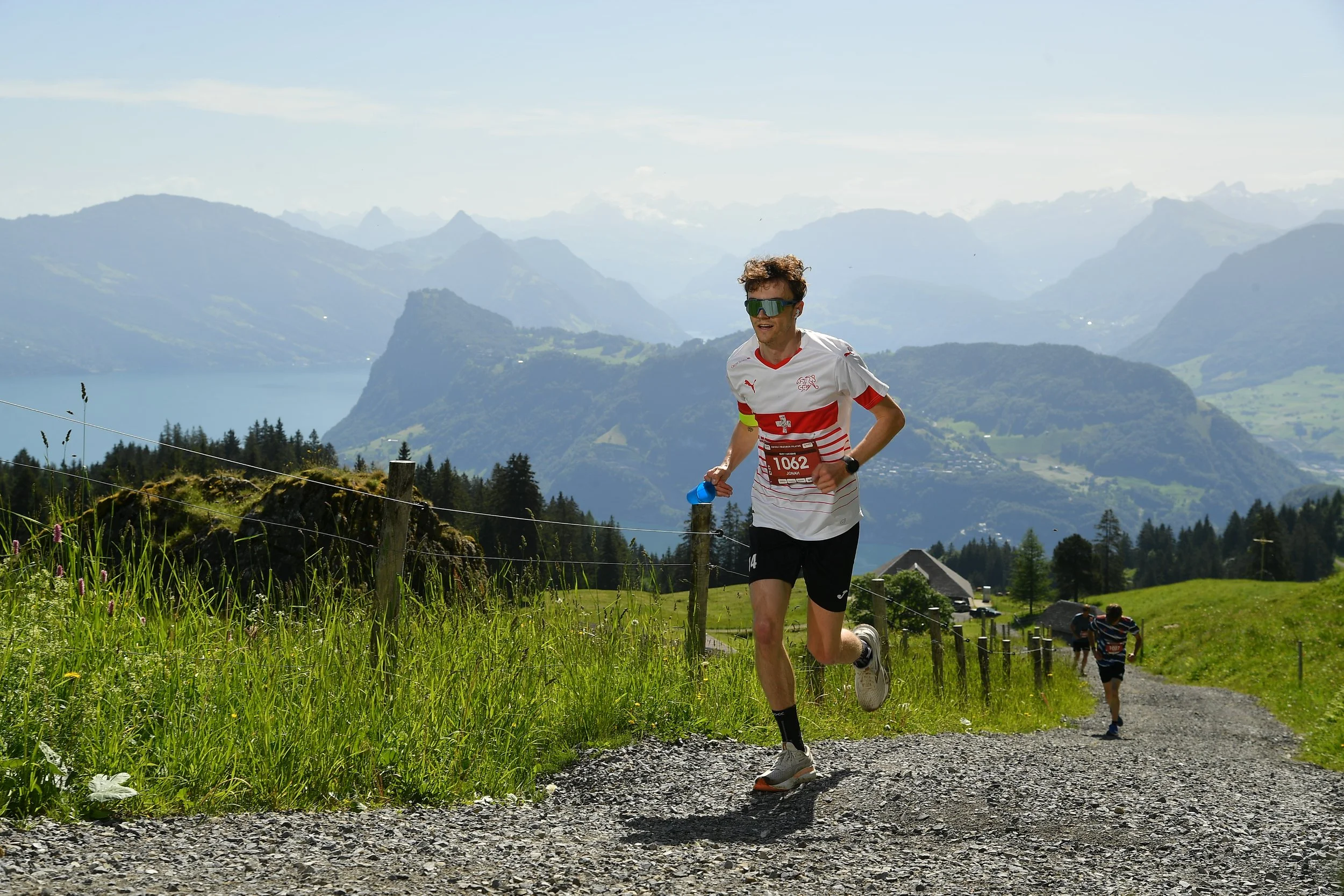 A man running outdoors on a gravel path with mountains in the background and a lake, wearing a white and red sports shirt, black shorts, sunglasses, and a watch.