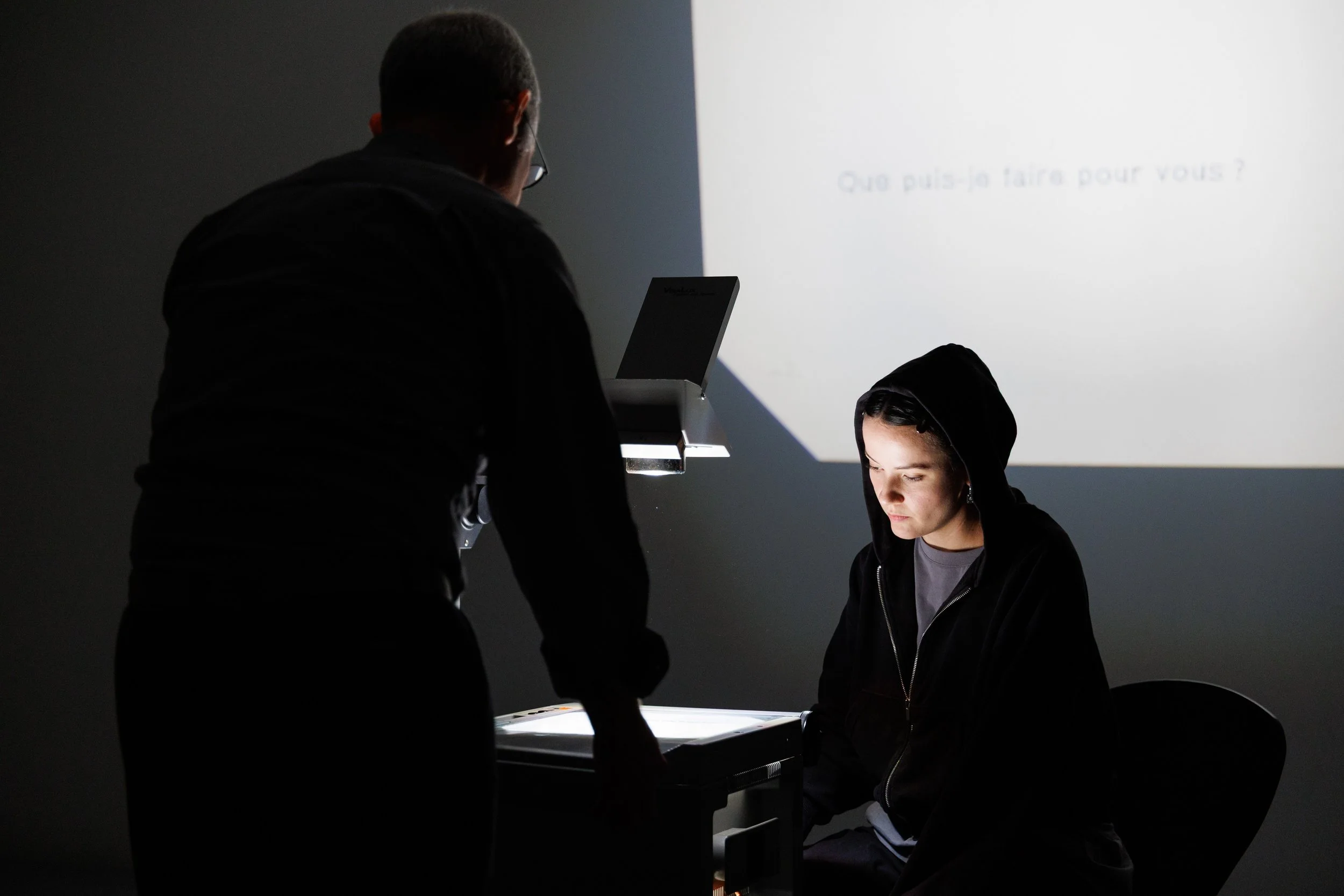 A person with dark hair and a hoodie sitting at a table, with another person standing beside him, in front of a white wall with writing in French. The lighting highlights their faces while the surroundings are dark.