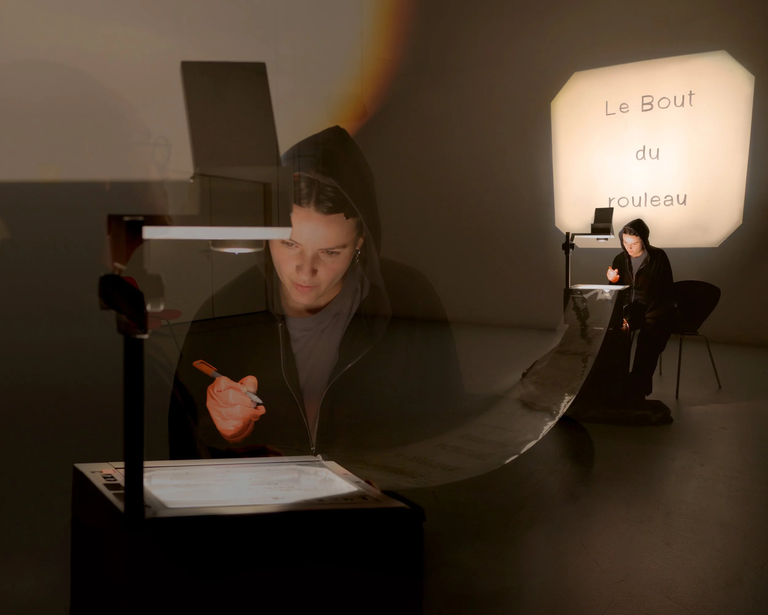 A woman with dark hair and a dark hoodie is sitting at a table with a music stand, writing on a paper. The setting appears to be an indoor room with a large, illuminated sign on the wall behind her that reads "Le Bout du rouleau". Reflection of the w