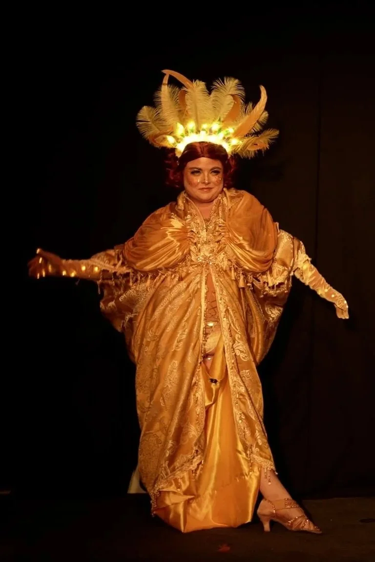 Person dressed in an elaborate gold costume with long sleeves, intricate embroidery, and a large feathered headdress emitting light, posing against a dark background.