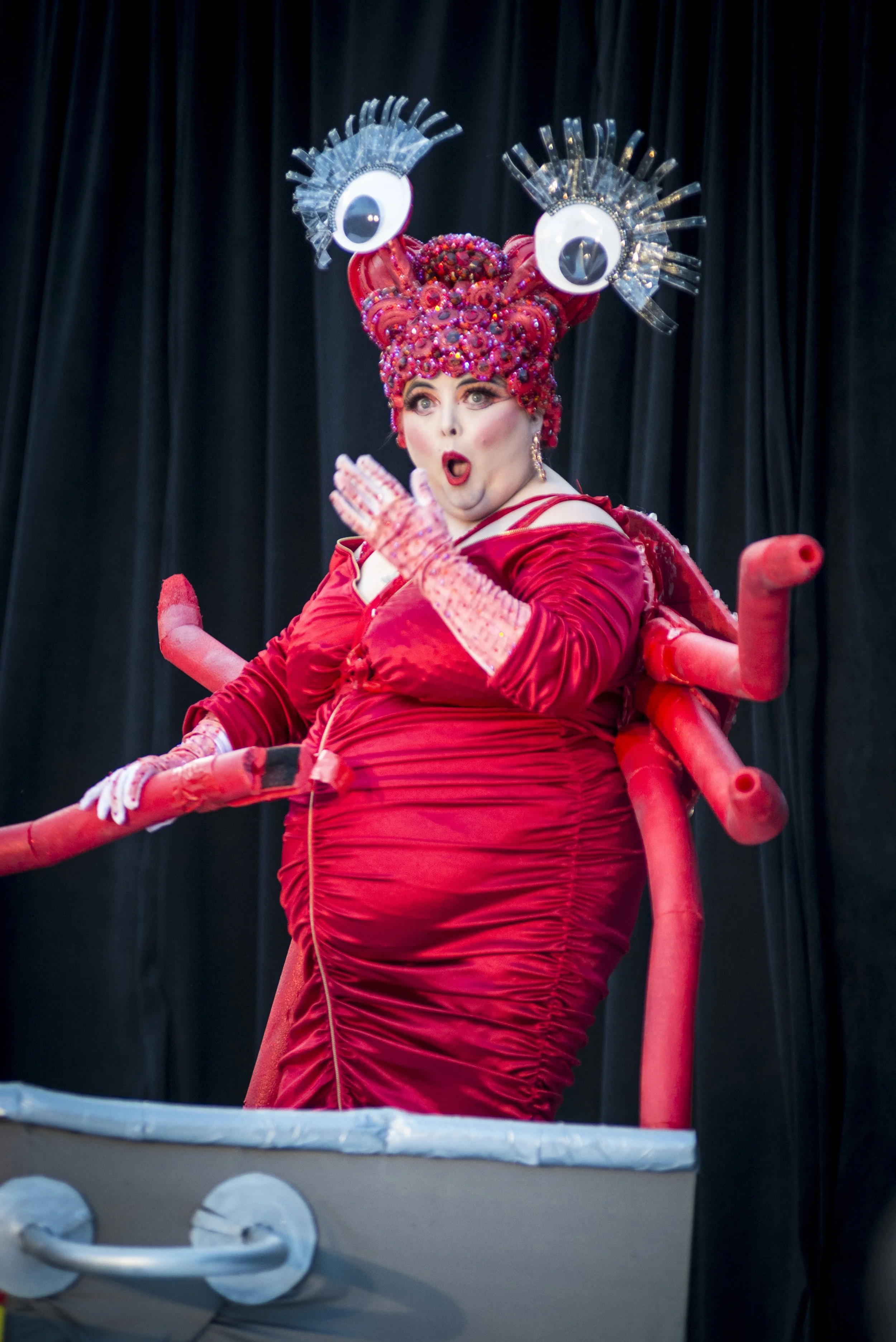 Performer dressed in a red costume with crab-like features, including large eyes and claw-like limbs, on stage with black curtain background.