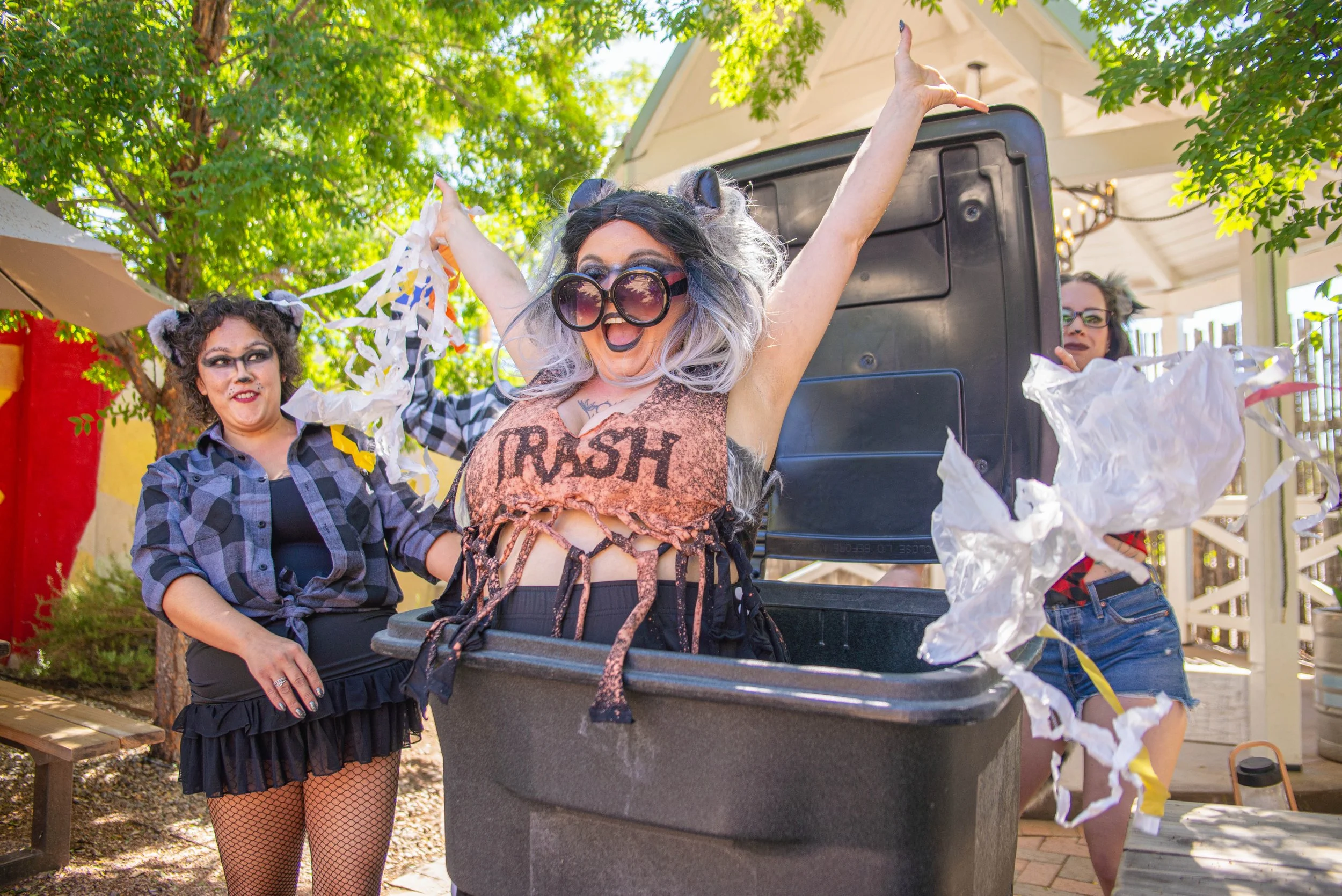 People in colorful costumes celebrating outdoors, with one woman jumping out of a trash bin with arms raised and others around her, confetti flying, trees and a house in the background.