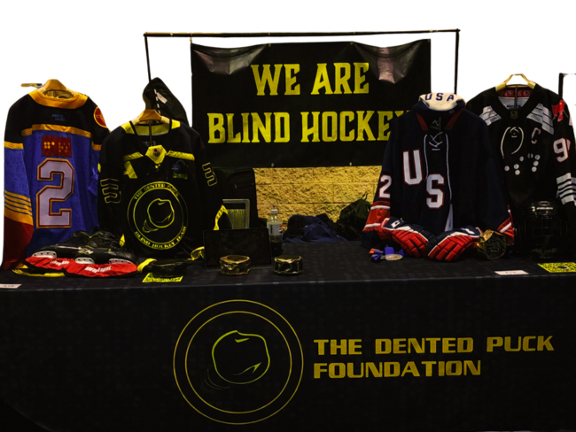 Display table at a blind hockey event featuring jerseys, a banner reading 'We Are Blind Hockey,' and materials from The Dented Puck Foundation.