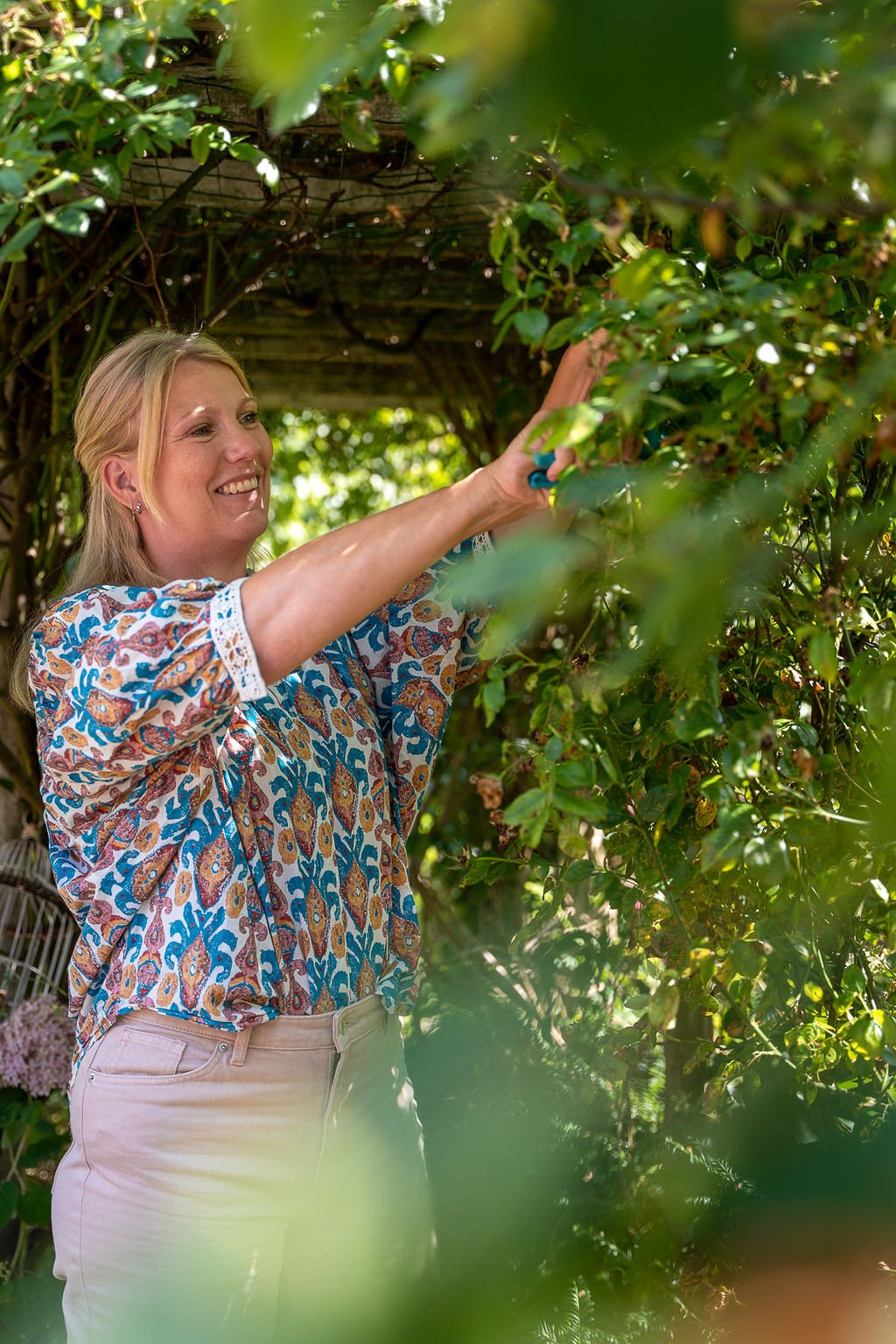 Vrouw plukker bloemen uit een groene struik in een tuin, glimlachend en in een kleurrijk blouse. - personal branding fotografie