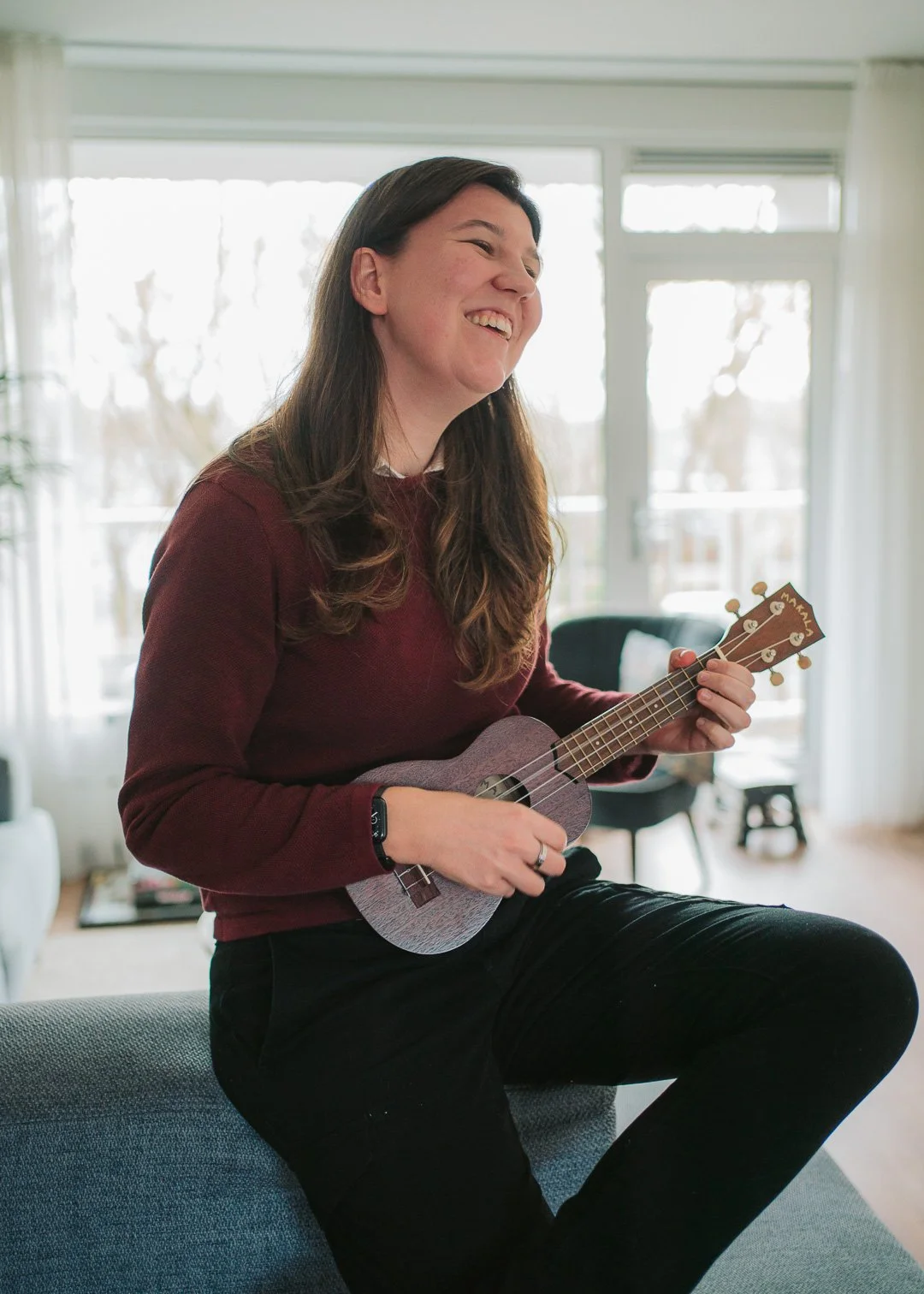Een vrouw met bruin haar speelt op een kleine ukelele, zittend op een bank in een lichte woonkamer met grote ramen.