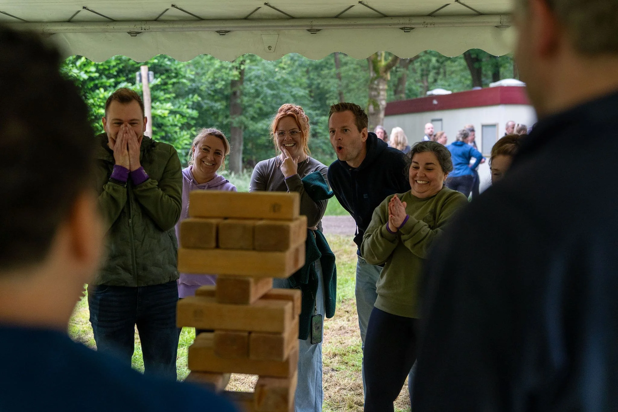 Mensen die enthousiast en lachend naar een groot Jenga-spel kijken onder een witte tent buiten, met een groene achtergrond van bomen. - evenementfotografie