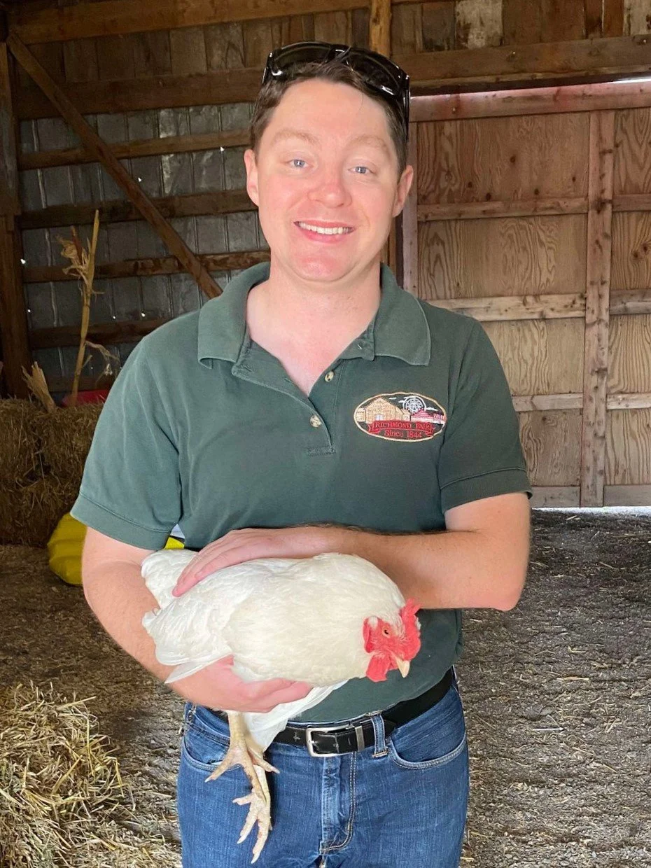 David Brown with sunglasses on his head, wearing a green polo shirt and blue jeans, holding a white chicken inside a wooden barn.