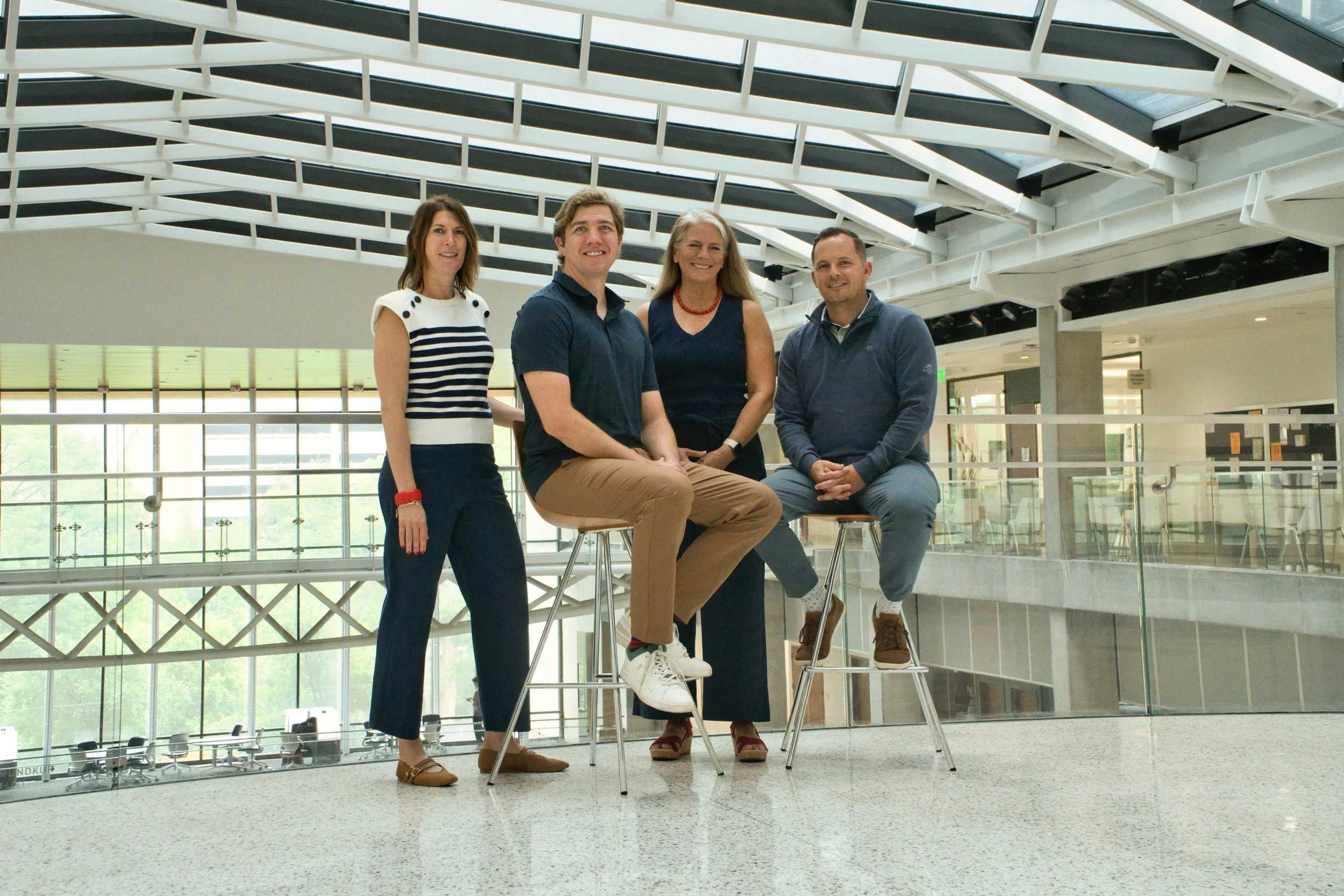 Four people, two women and two men, sitting and standing on stools in an indoor space with large windows and glass railing, under a modern glass ceiling.