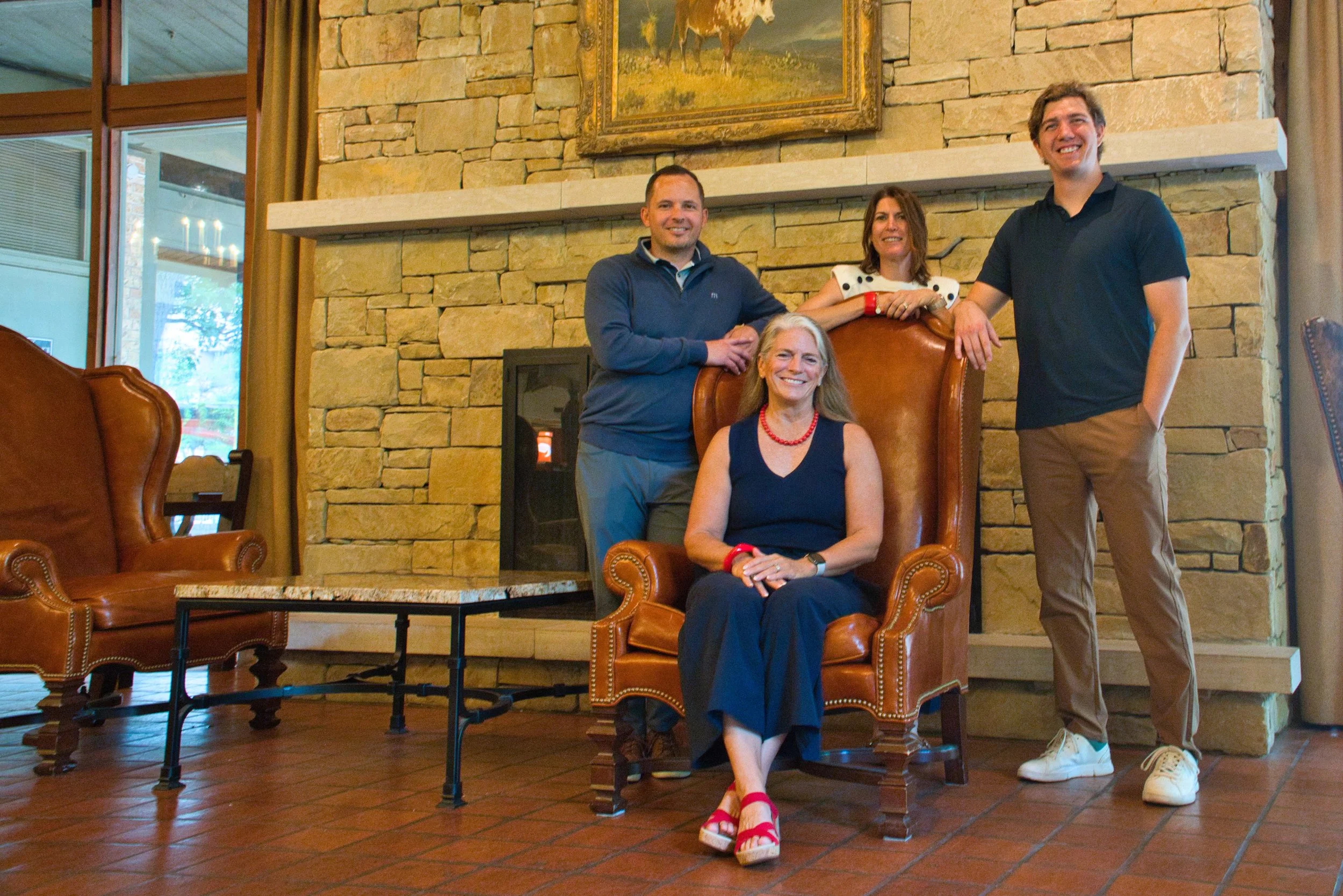 Four people, three men and one woman, smiling and posing in a warmly lit room with a stone fireplace. The woman is seated in a large leather chair, wearing a dark blue dress and red accessories. Two men stand behind her, one on each side, and another woman stands behind the seated woman, leaning on the chair's back.