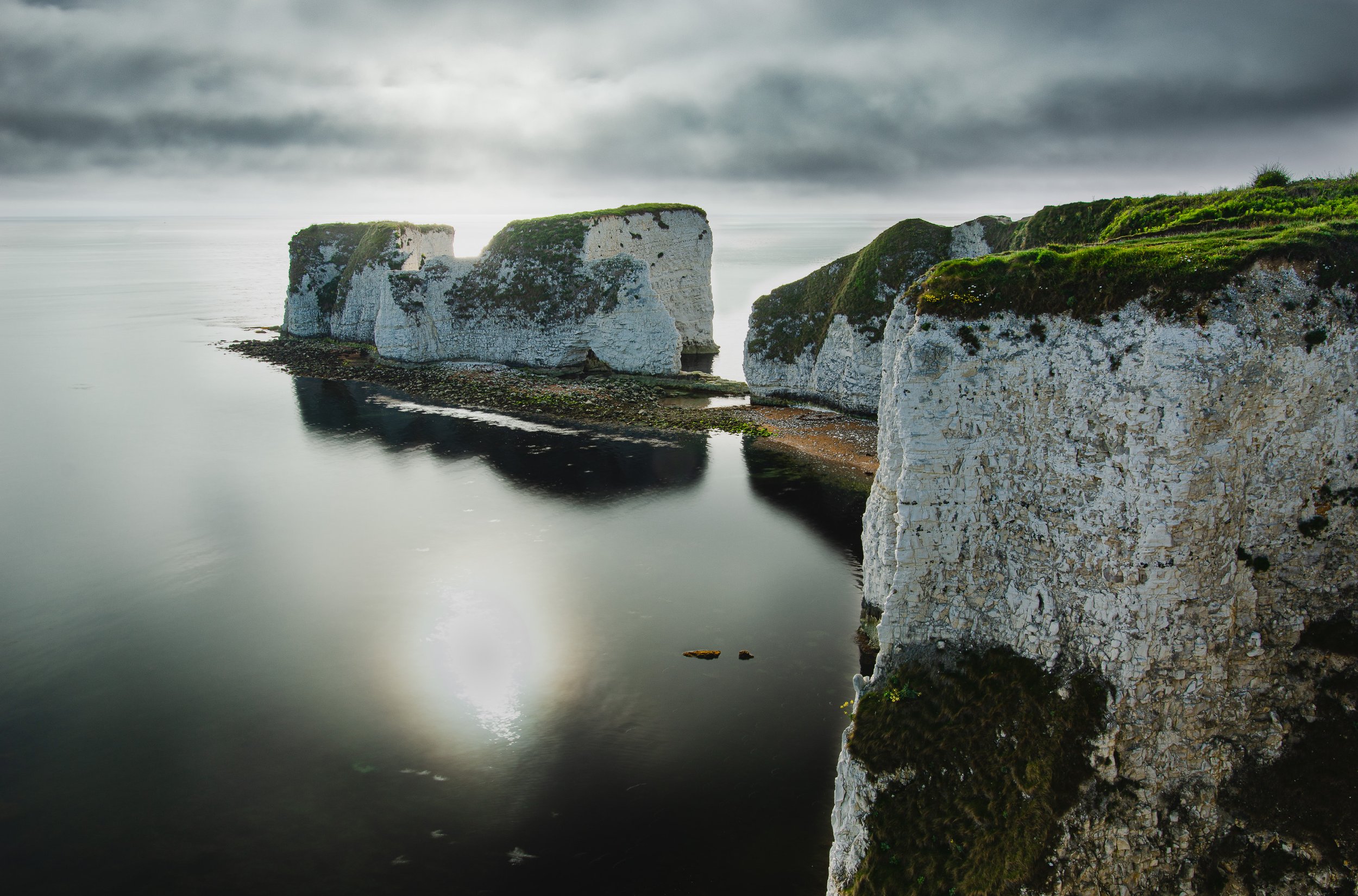 White chalk cliffs along the coastline with green grass on top, reflected in calm water, with dark, cloudy sky overhead.