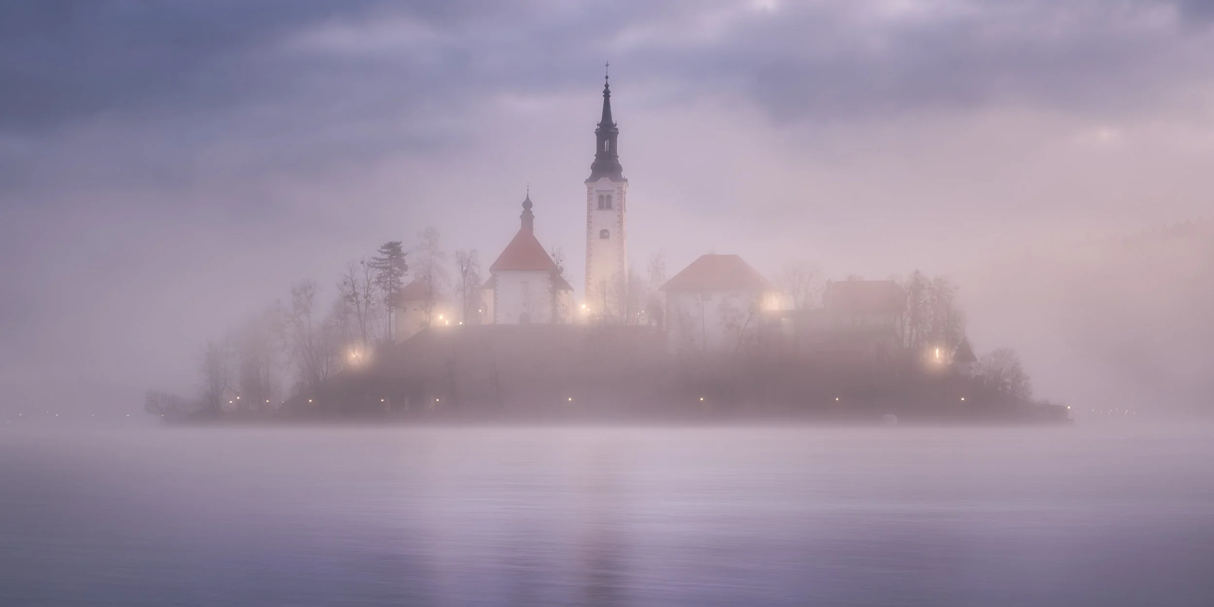 A church with a tall steeple, situated on a hill surrounded by trees, on a foggy day over a body of water.