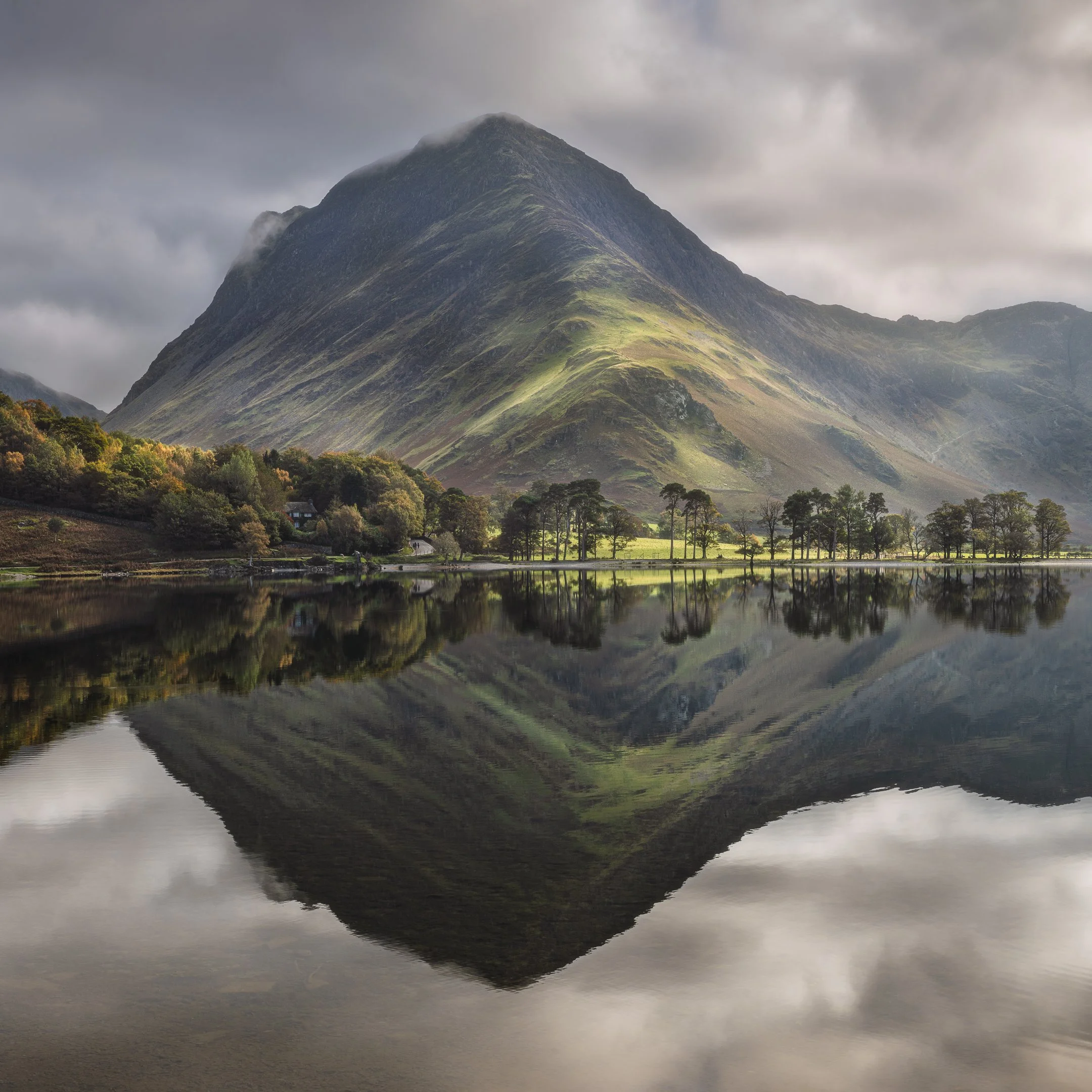 A tall mountain with steep slopes and patches of green grass, partly shrouded in fog, reflected in a calm body of water at the base, with a cloudy sky overhead.