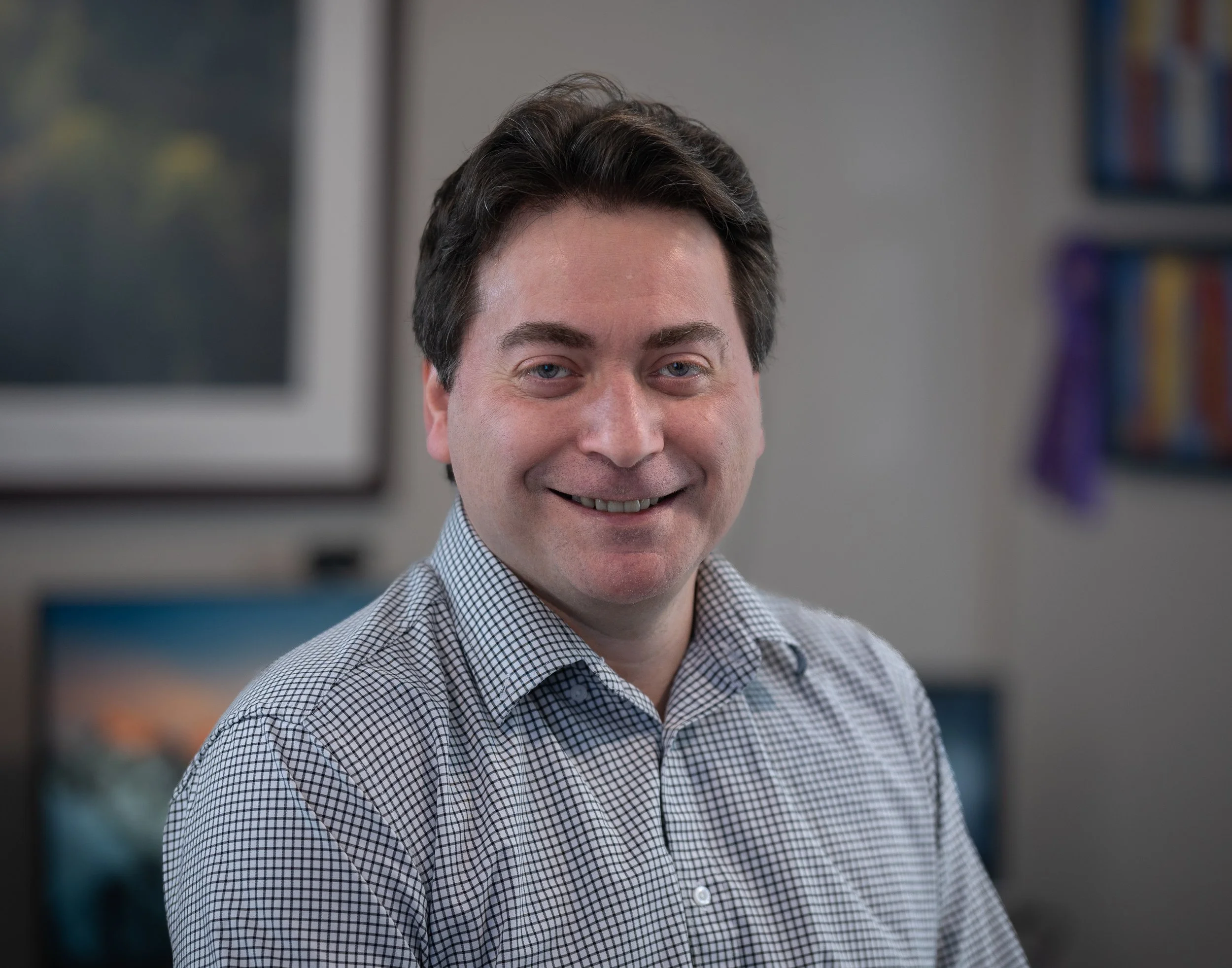 A smiling man with light skin, dark brown hair, and blue eyes, wearing a checkered shirt, appears to be in an office or professional setting with blurred background including bookshelves.
