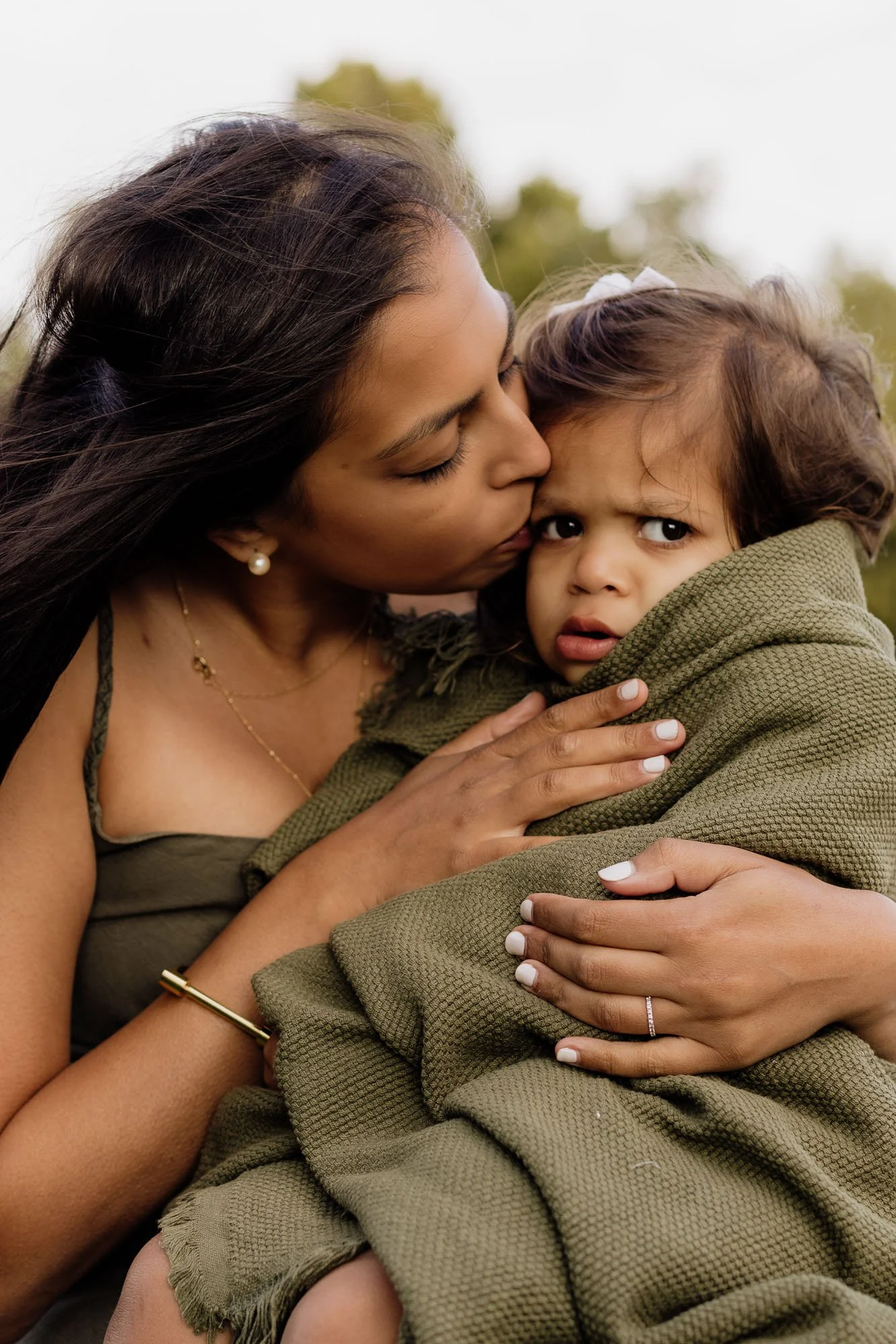 Mum hugs her toddler daughter tight, wrapped in a picnic blanket, and kisses her on the head