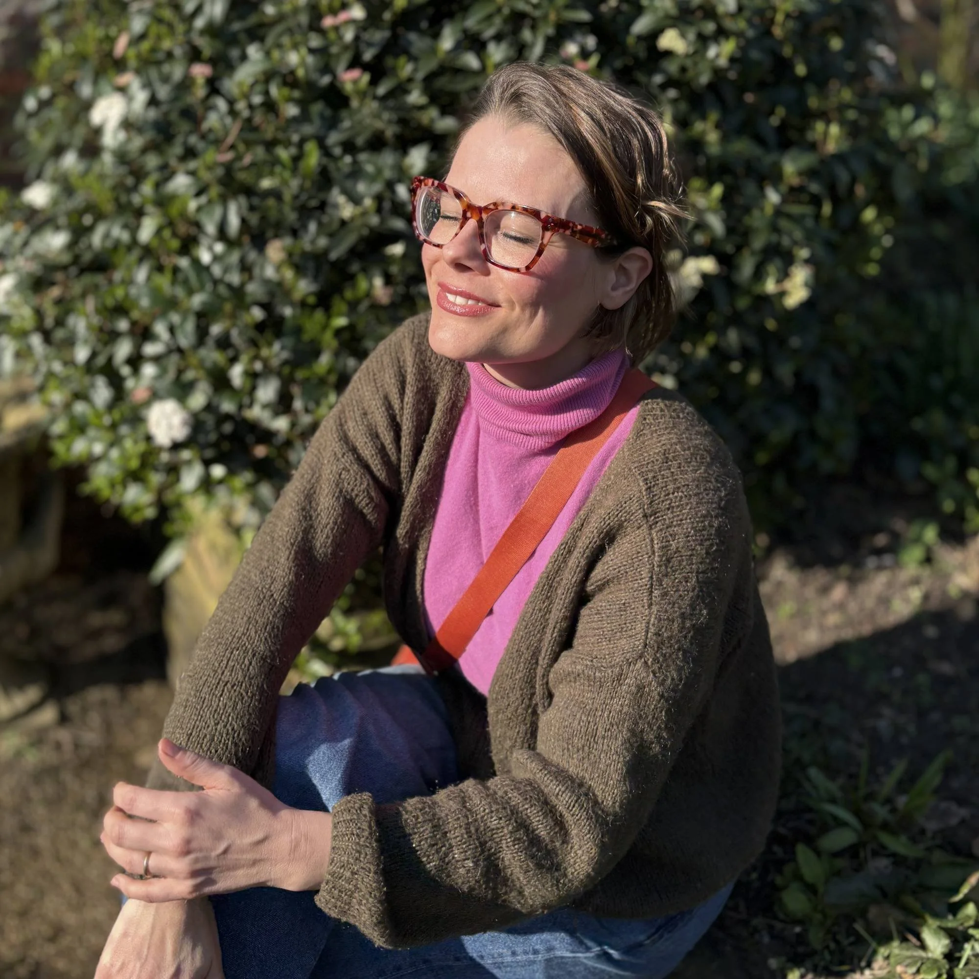 Artist and Illustrator Emma Winterschladen, wearing tortoiseshell glasses, a pink turtleneck, and a brown sweater, with her eyes closed, smiling outdoors in sunlight, against a green leafy background.