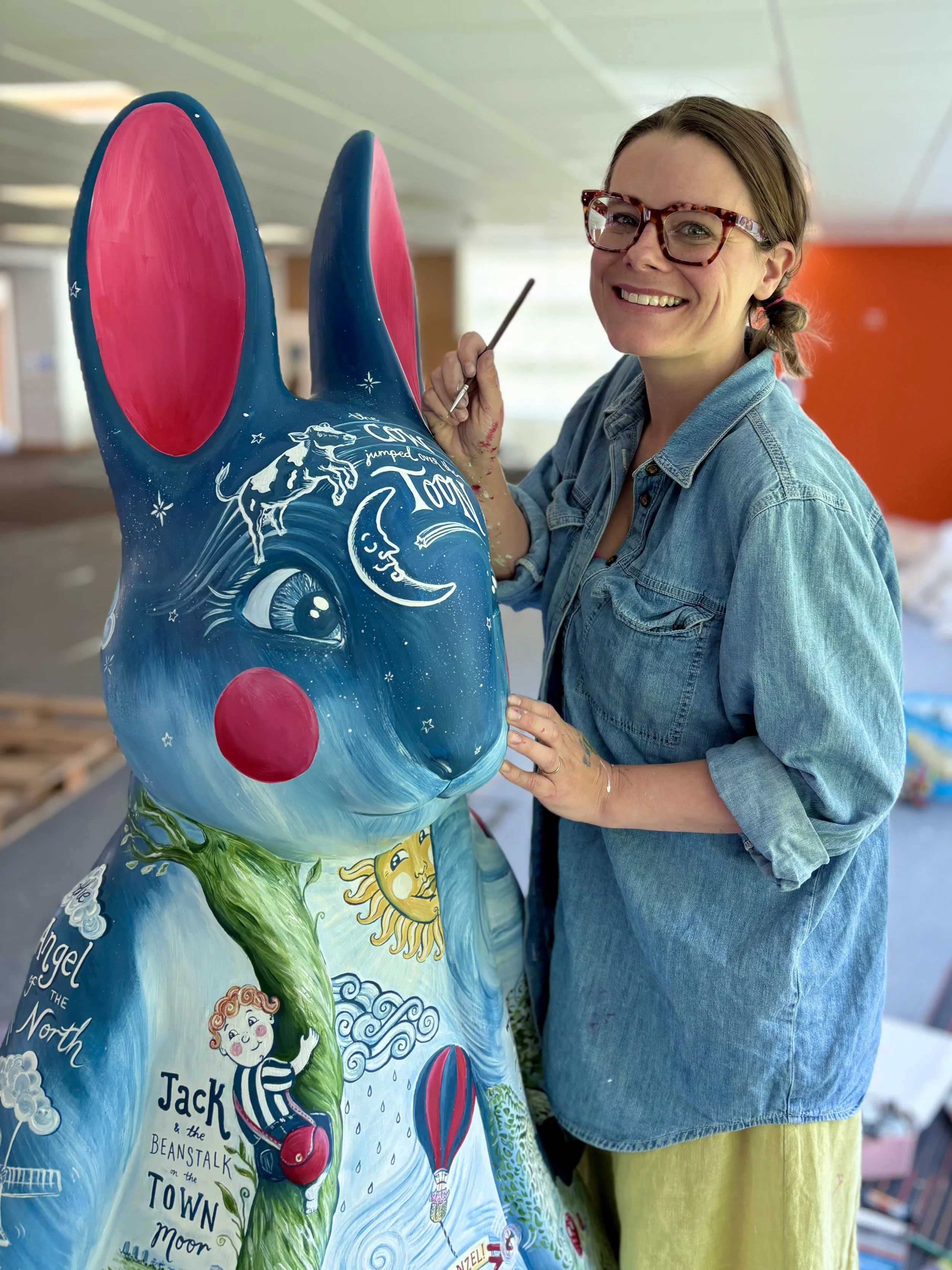 Emma Winterschladen, an artist and illustrator wearing glasses and a denim shirt painting a a whimsical Peter Rabbit sculpture, featuring fairytale characters, animals, and celestial elements.