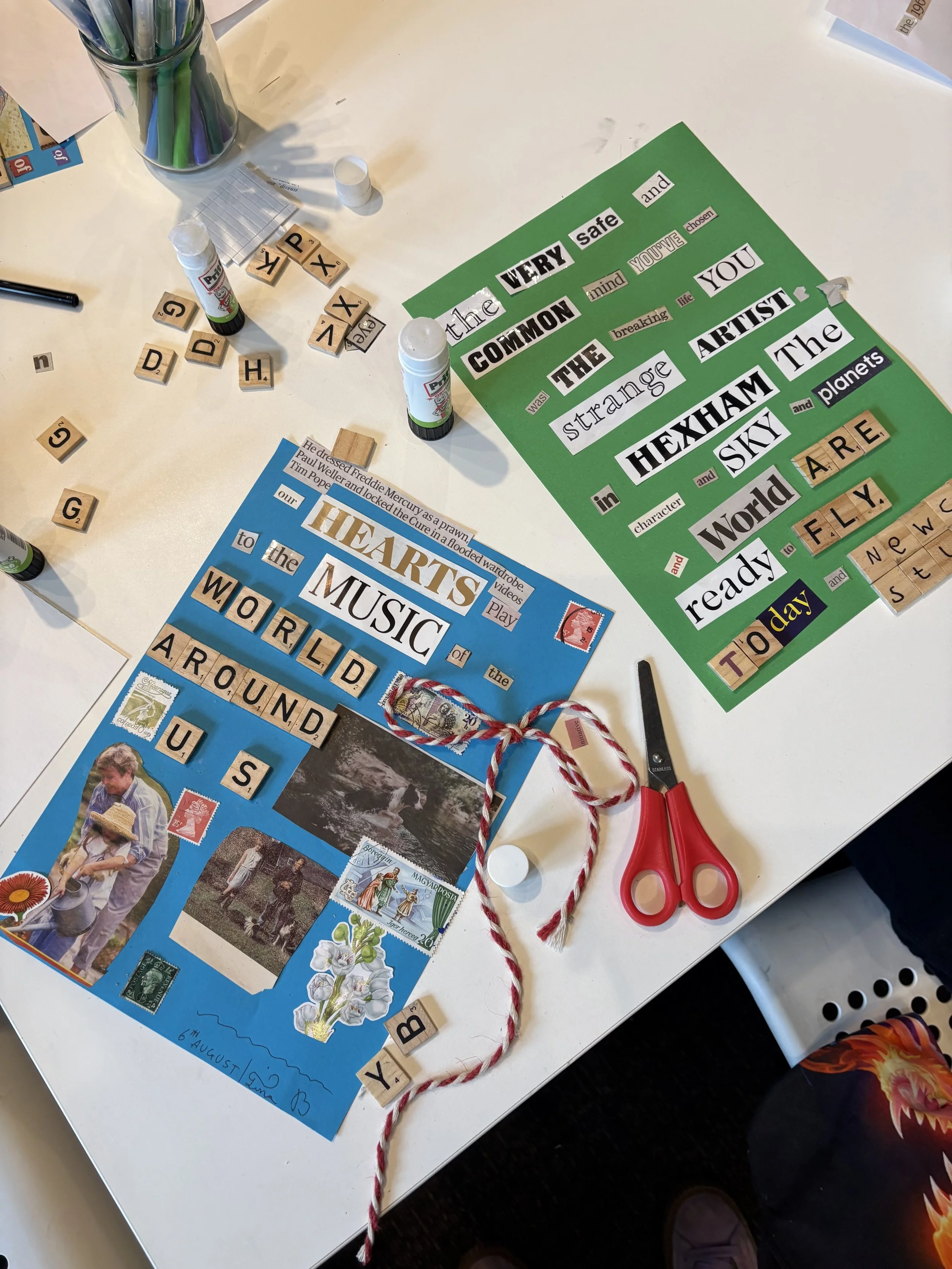 A craft project on a white table with a green poster that has cut-out words and phrases about art and creativity, a pair of red scissors, glue stick, twisted red and white string, and various stamps and images, including a photo of children outdoors, a butterfly, and postage stamps.