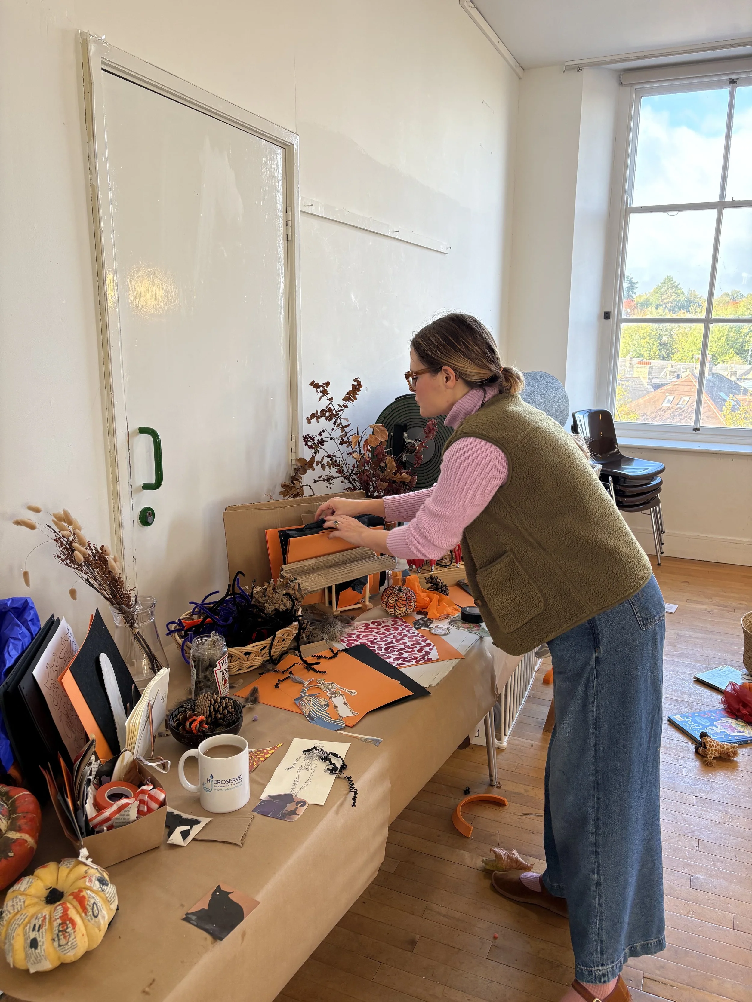 Artist Emma Winterschladen hosting a halloween workshop, wearing glasses, a green vest and lilac sweater, is standing at a table filled with arts and crafts supplies in a room with large window and wooden floor.