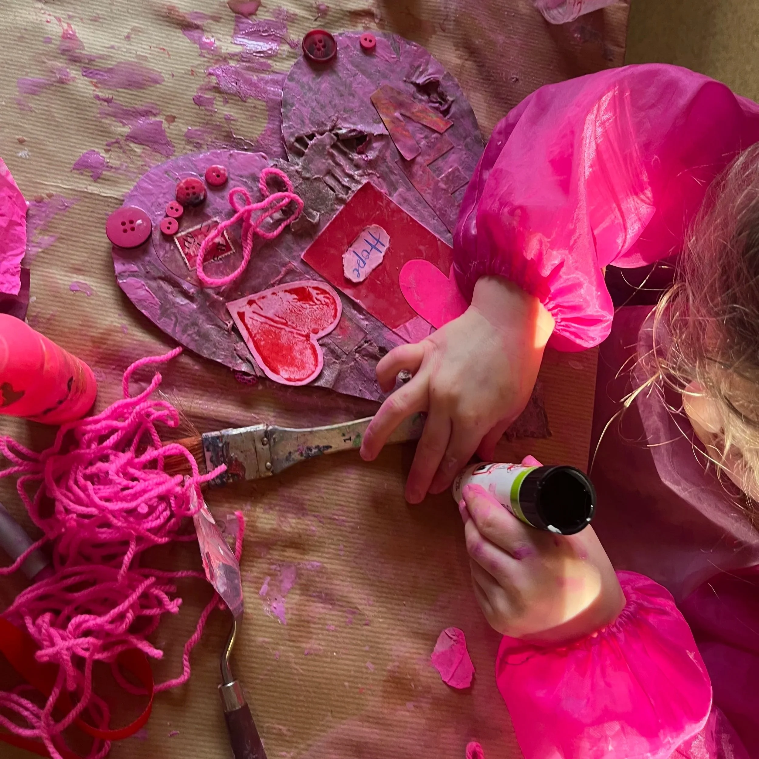 A table with a craft project consisting of a large pink paper heart, pink yarn, buttons, and scraps of pink tissue paper, with a child's hand working on the craft.
